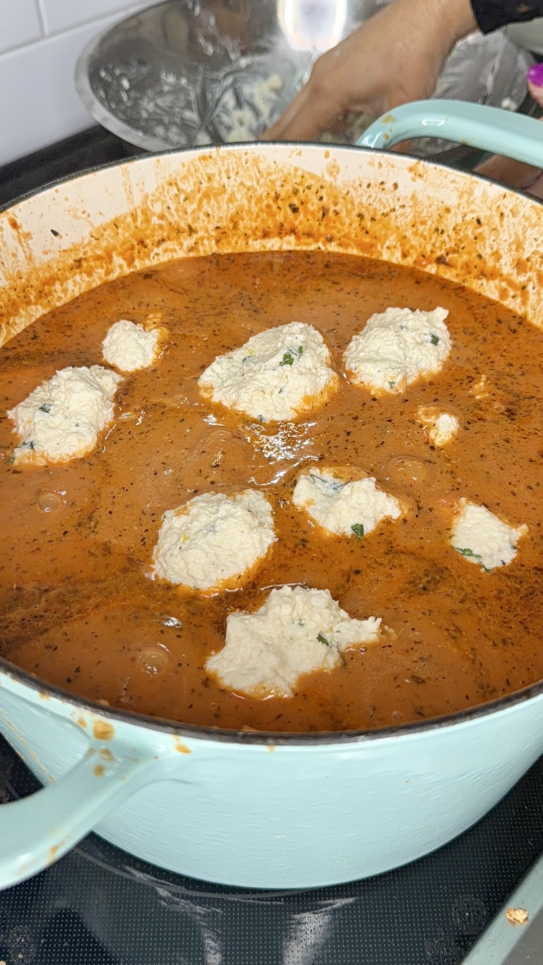 A light blue pot filled with rich, red tomato sauce and several white ricotta cheese dollops on top. A person’s hand is visible in the background mixing ingredients in a bowl.