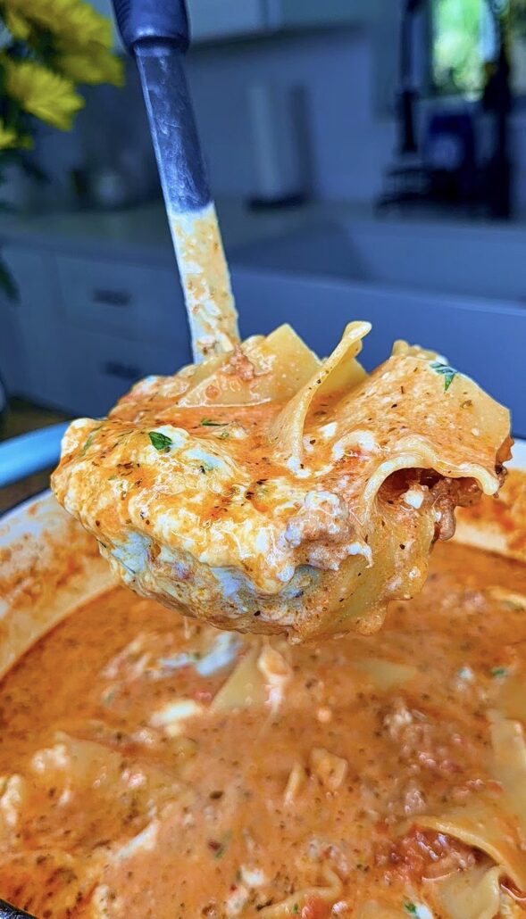 A close-up of a ladle lifting a serving of creamy lasagna soup with lasagna noodles, cheese, and herbs from a pot filled with rich, tomato-based broth. Kitchen counters and cabinets appear in the blurred background.