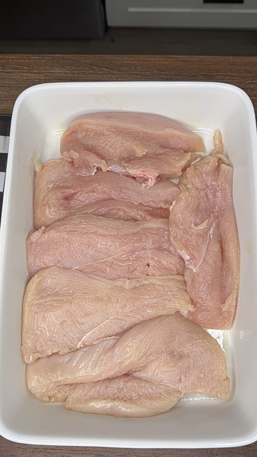 Raw chicken breast fillets arranged in a white rectangular dish, ready for cooking, on a wooden countertop.