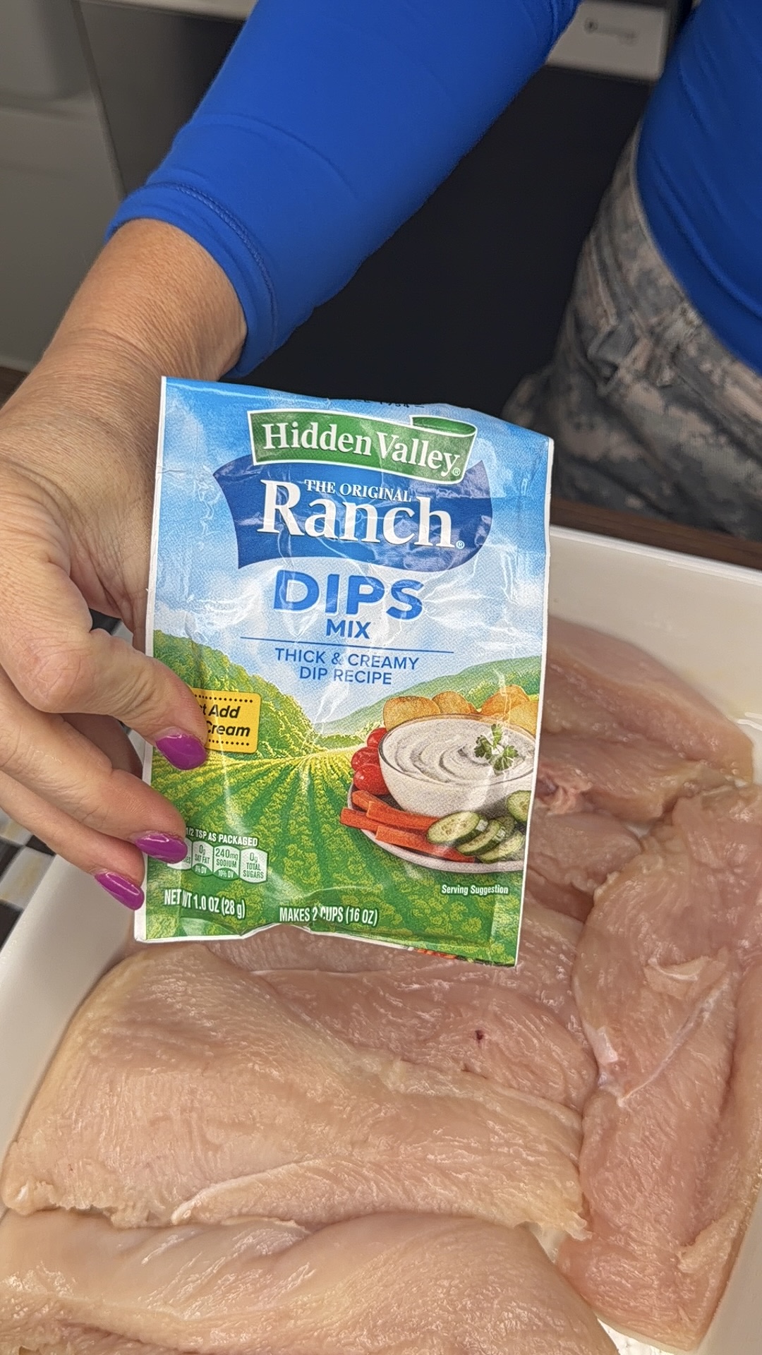 A person with pink nails holds a packet of Hidden Valley Ranch Dip Mix over a dish filled with raw chicken breasts.