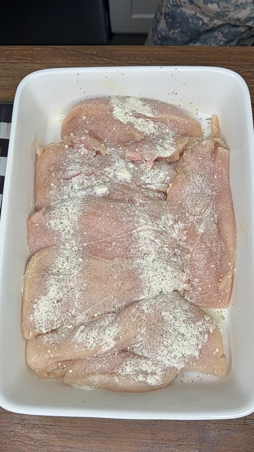 Raw chicken breasts arranged in a white rectangular baking dish, sprinkled with a layer of seasoning and spices on top, sitting on a wooden surface.