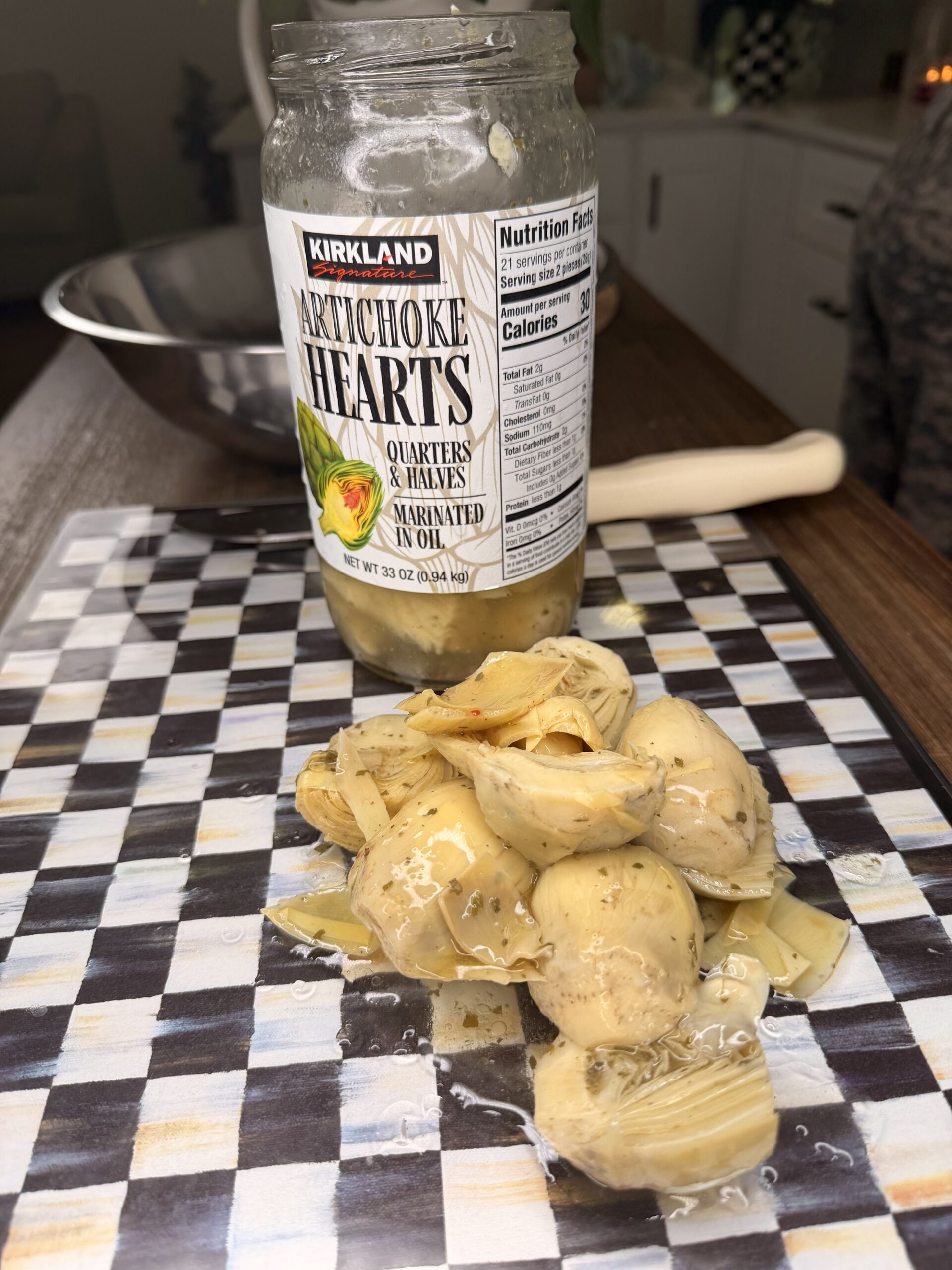 A jar of Kirkland Artichoke Hearts, marinated in oil, sits open on a checkered cutting board with artichoke hearts spilled out in front of it. A kitchen bowl and utensils are visible in the background.