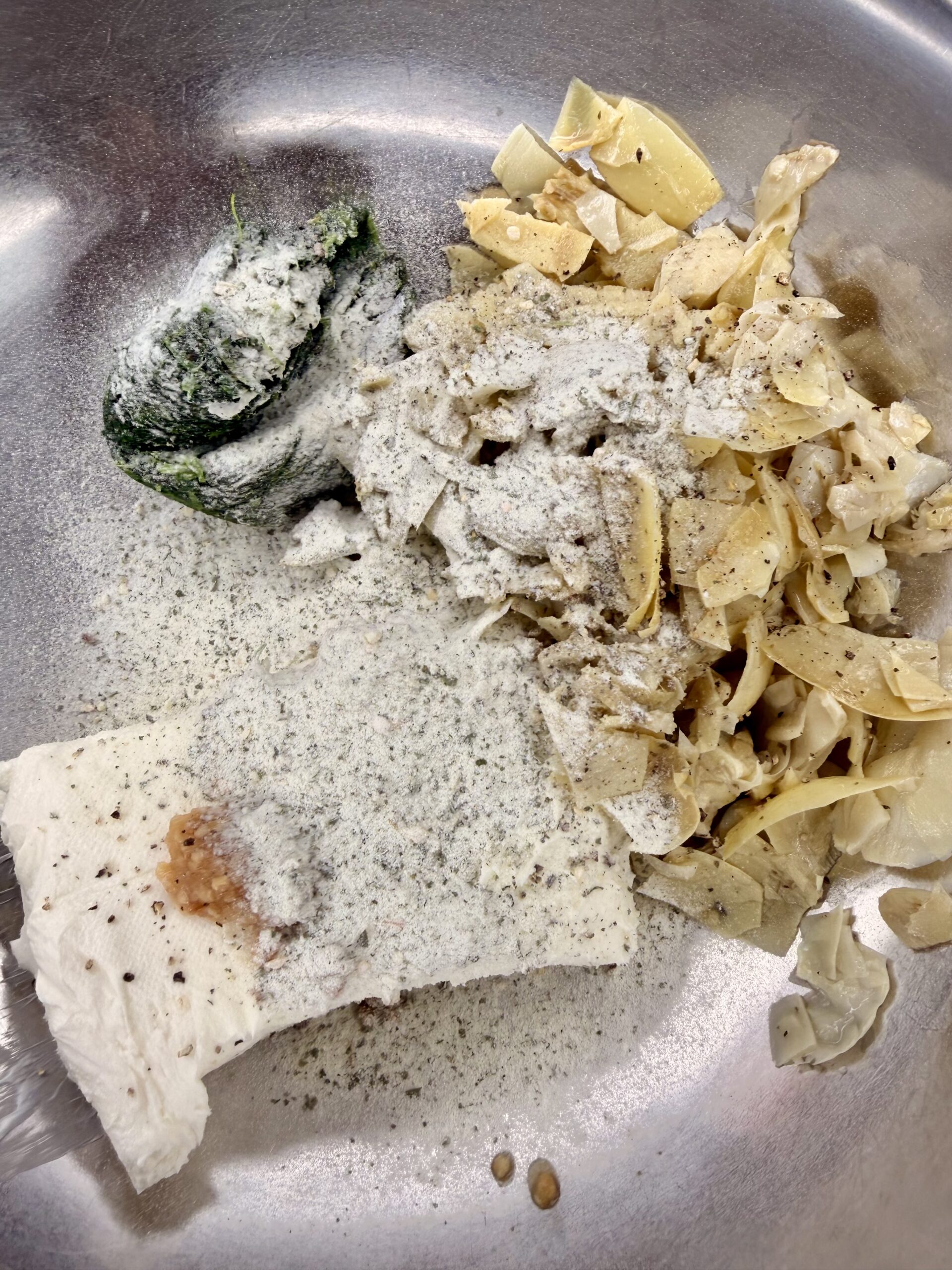 A metal bowl containing a block of cream cheese, cooked egg noodles, a lump of chopped green spinach, and various white and black powdered seasonings sprinkled on top.