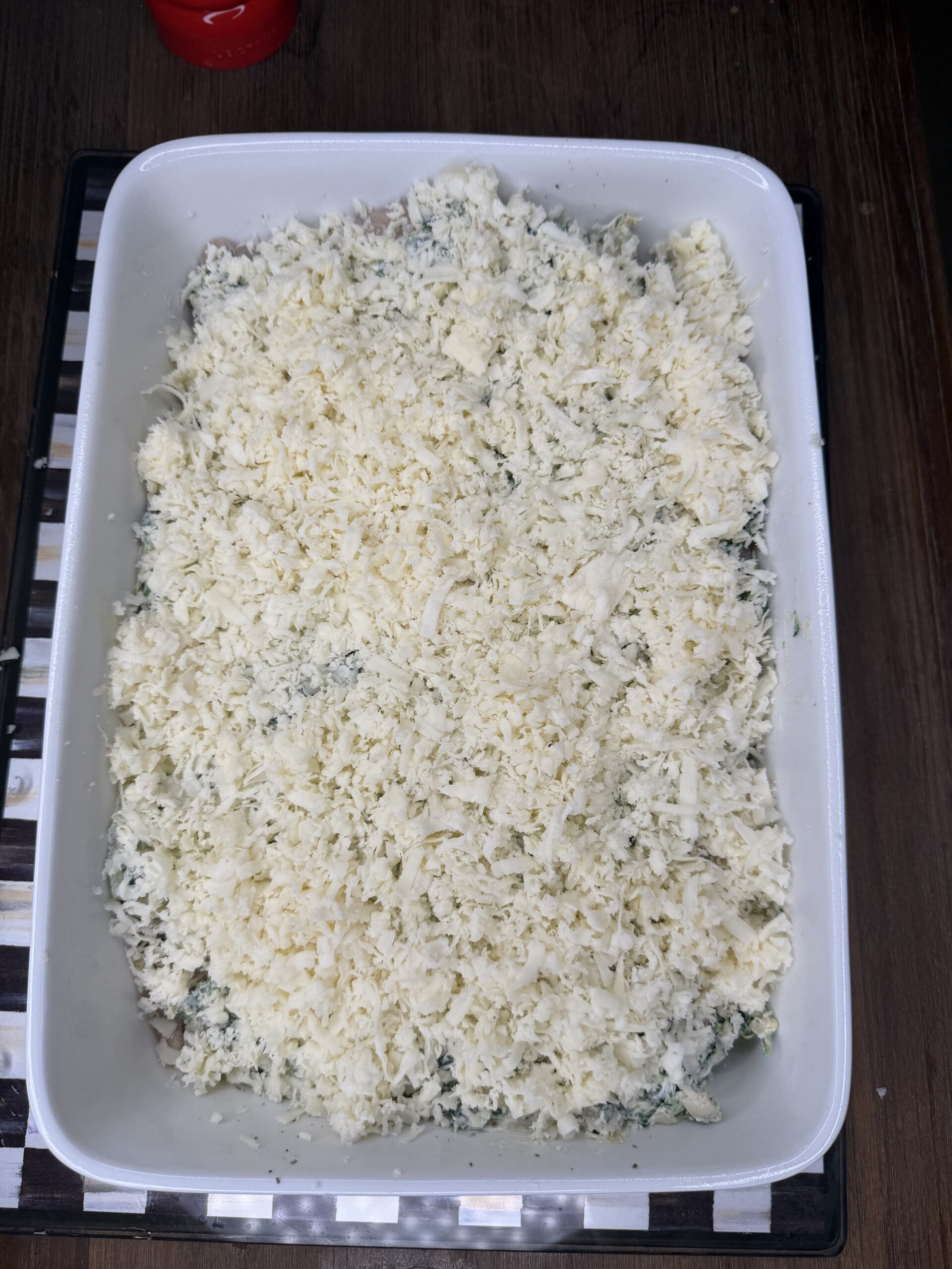 A white baking dish filled with a casserole topped with a generous layer of shredded cheese, ready to be baked, sits on a dark table. The casserole appears creamy with green herbs visible underneath the cheese.