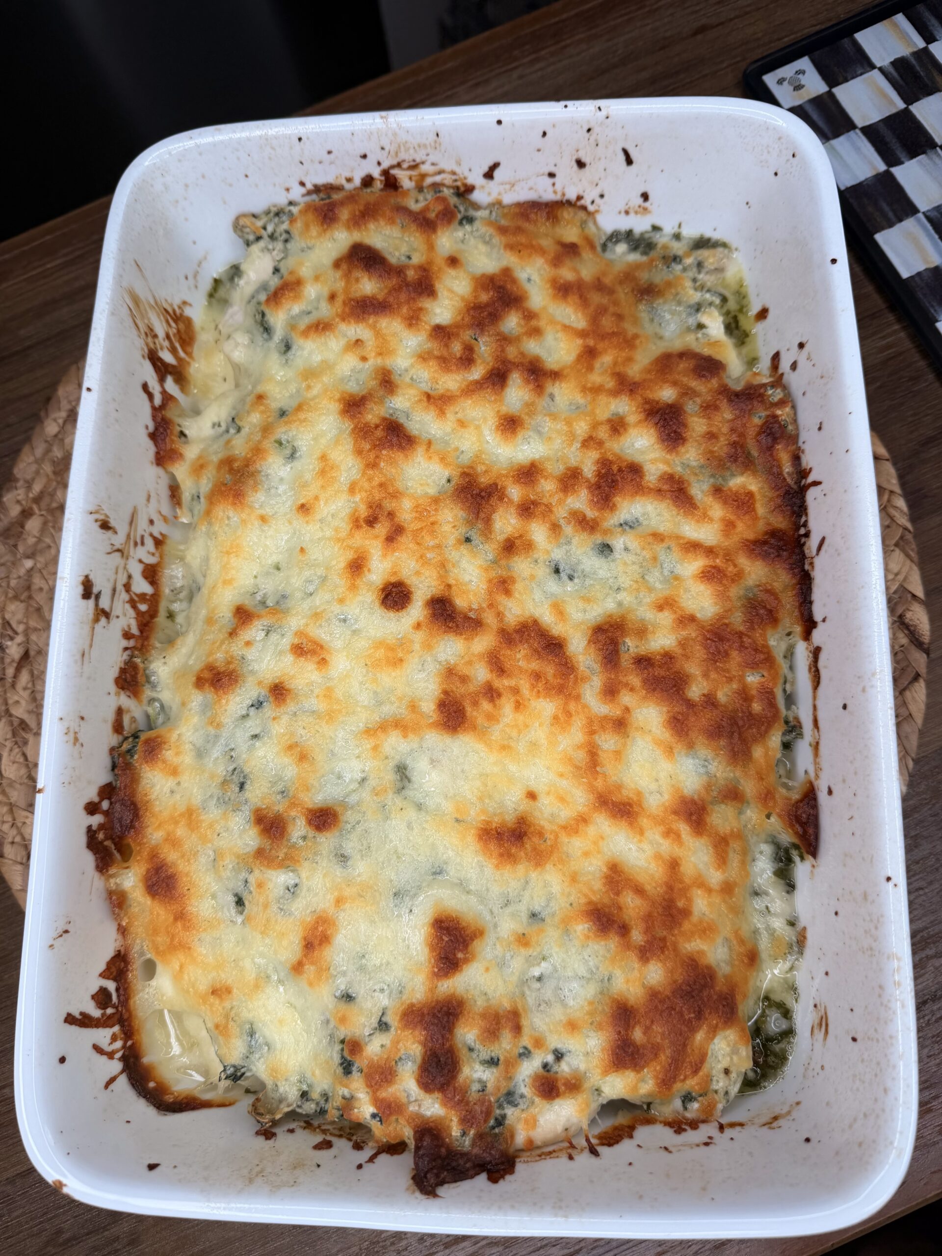 A white baking dish filled with baked lasagna topped with melted, golden-brown cheese sits on a wooden surface. The edges of the cheese are slightly crispy and browned.