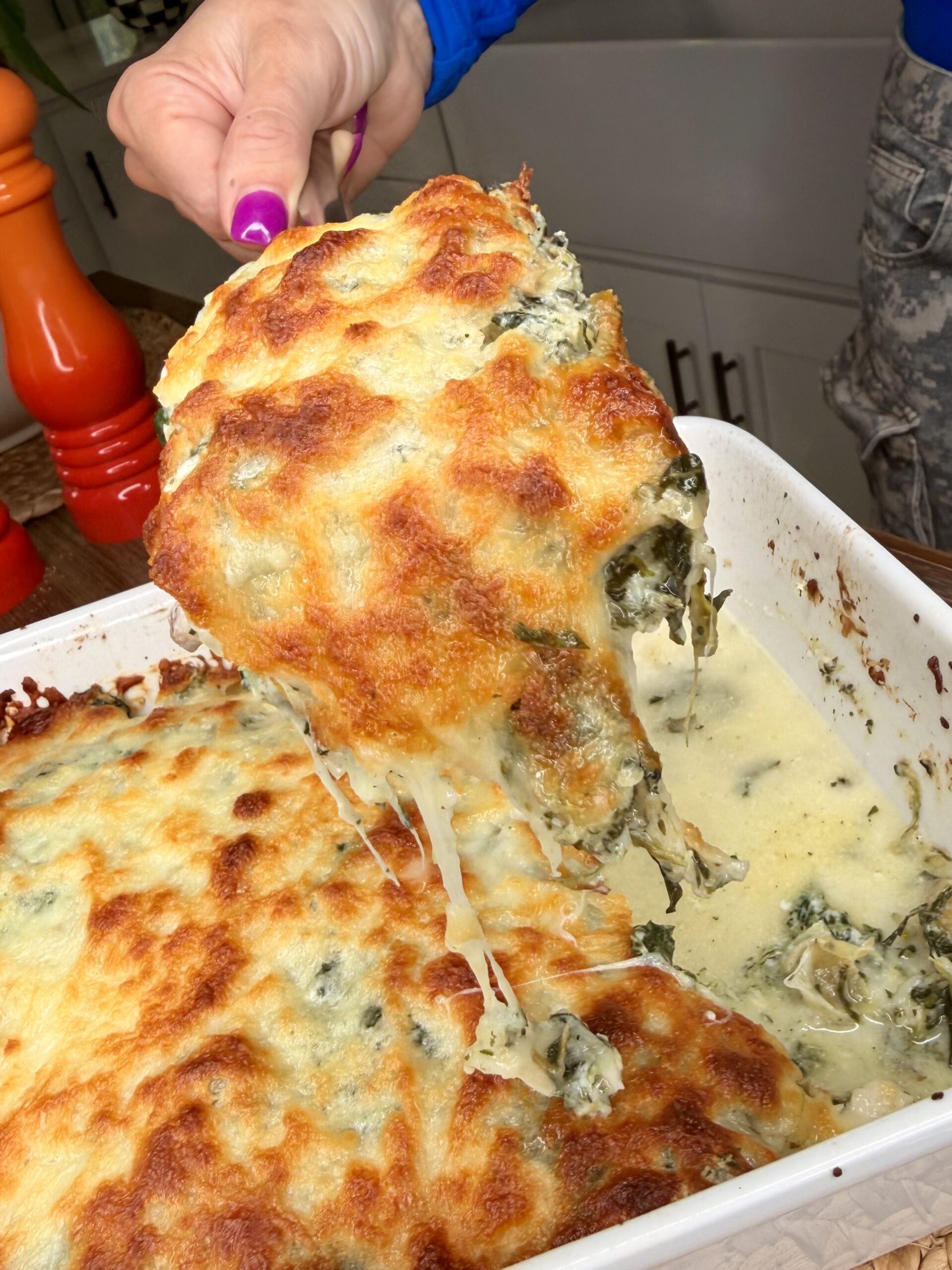 A hand holds a large serving of cheesy spinach casserole with melted, golden-brown cheese stretching from a white baking dish.