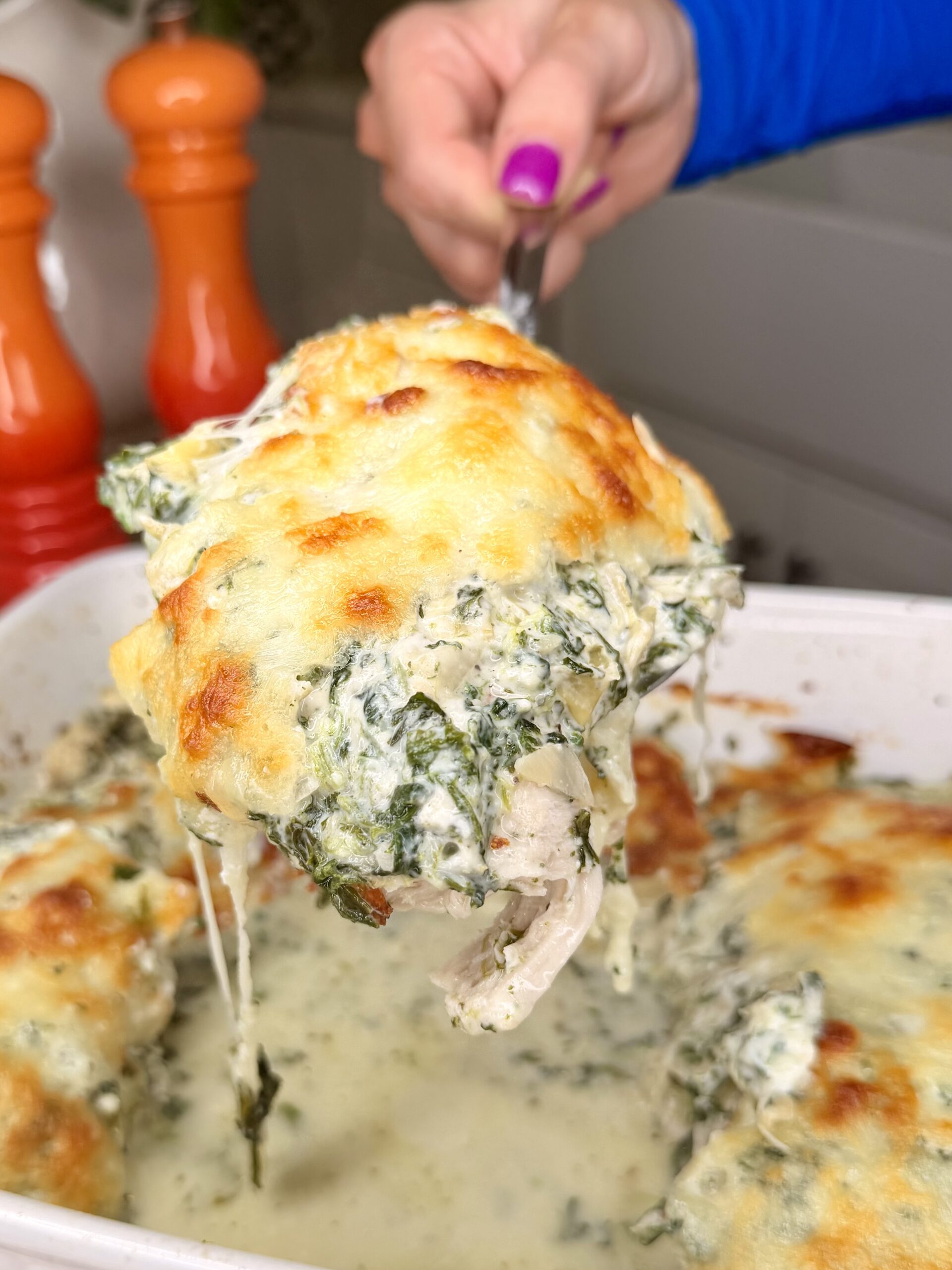 A hand holds a serving utensil lifting a cheesy, baked chicken breast topped with creamy spinach from a casserole dish. Melted cheese stretches, and orange pepper grinders are seen in the background.