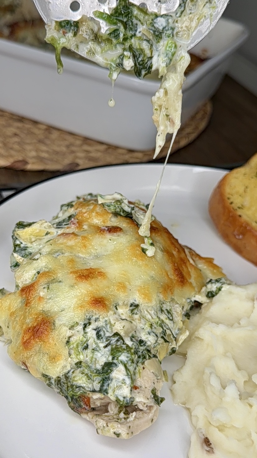 A serving of creamy baked chicken topped with melted cheese and spinach is being lifted onto a plate with mashed potatoes and a slice of garlic bread. Cheese is stretching from the main dish.