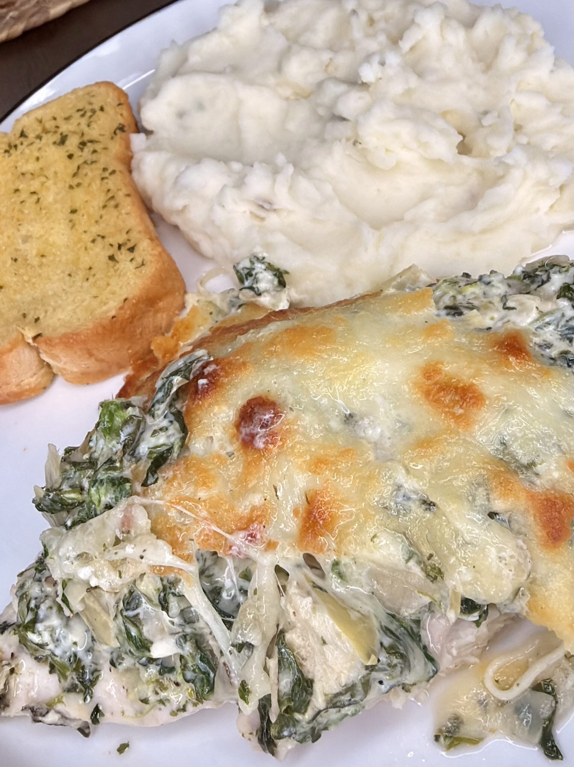 A plate with creamy mashed potatoes, a slice of garlic bread, and a portion of baked chicken topped with melted cheese, spinach, and a creamy sauce.