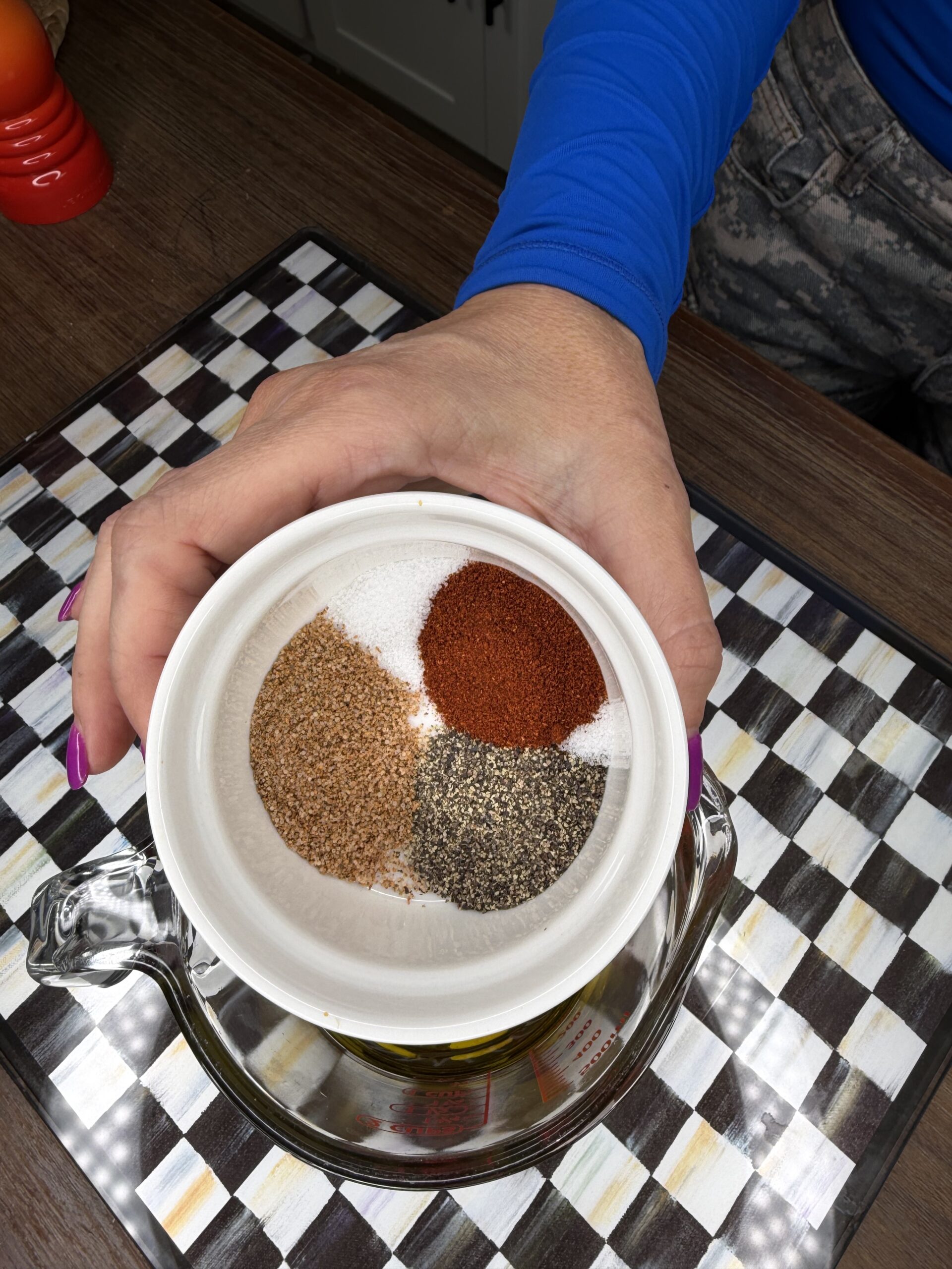 A hand with purple nails holds a small white bowl containing four spices—salt, paprika, black pepper, and brown seasoning—over a glass container on a checkered surface.