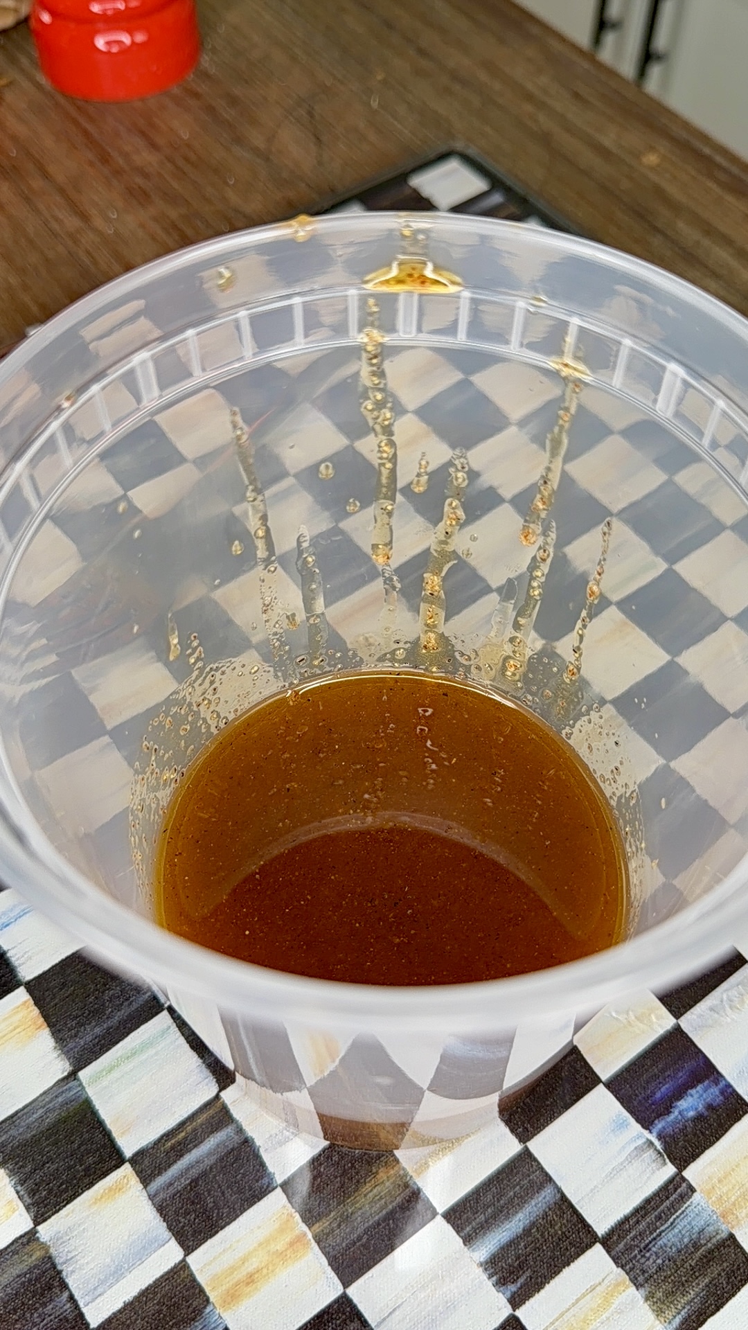 A clear plastic container partially filled with a brown liquid sits on a checkered-patterned surface. Some of the liquid has splashed onto the sides of the container.