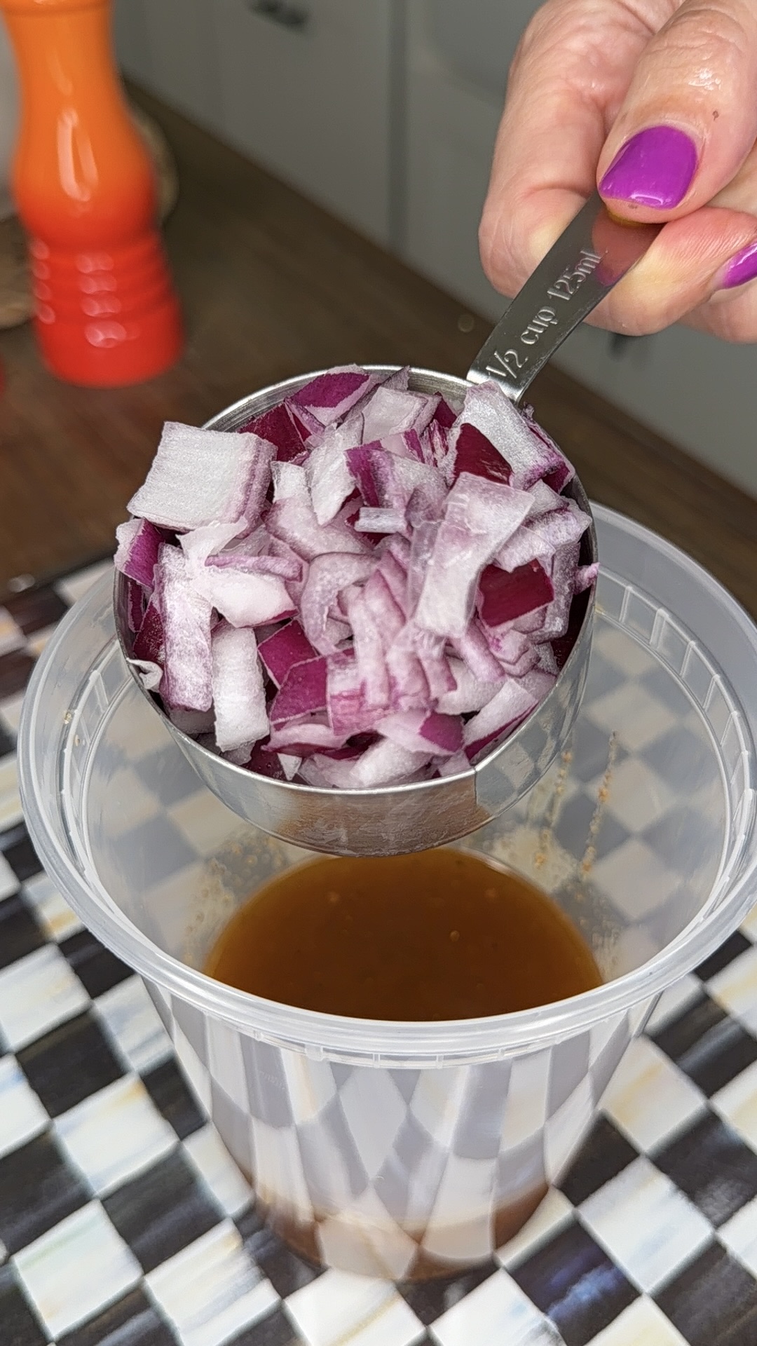 A hand with pink nail polish holds a metal half-cup filled with chopped red onions over a plastic container with brown liquid, on a checkered surface.