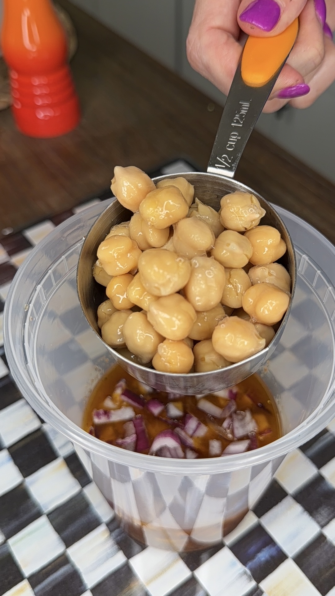 A hand with purple nail polish holds a metal measuring cup of chickpeas above a plastic container with chopped red onions and liquid on a checkered surface.