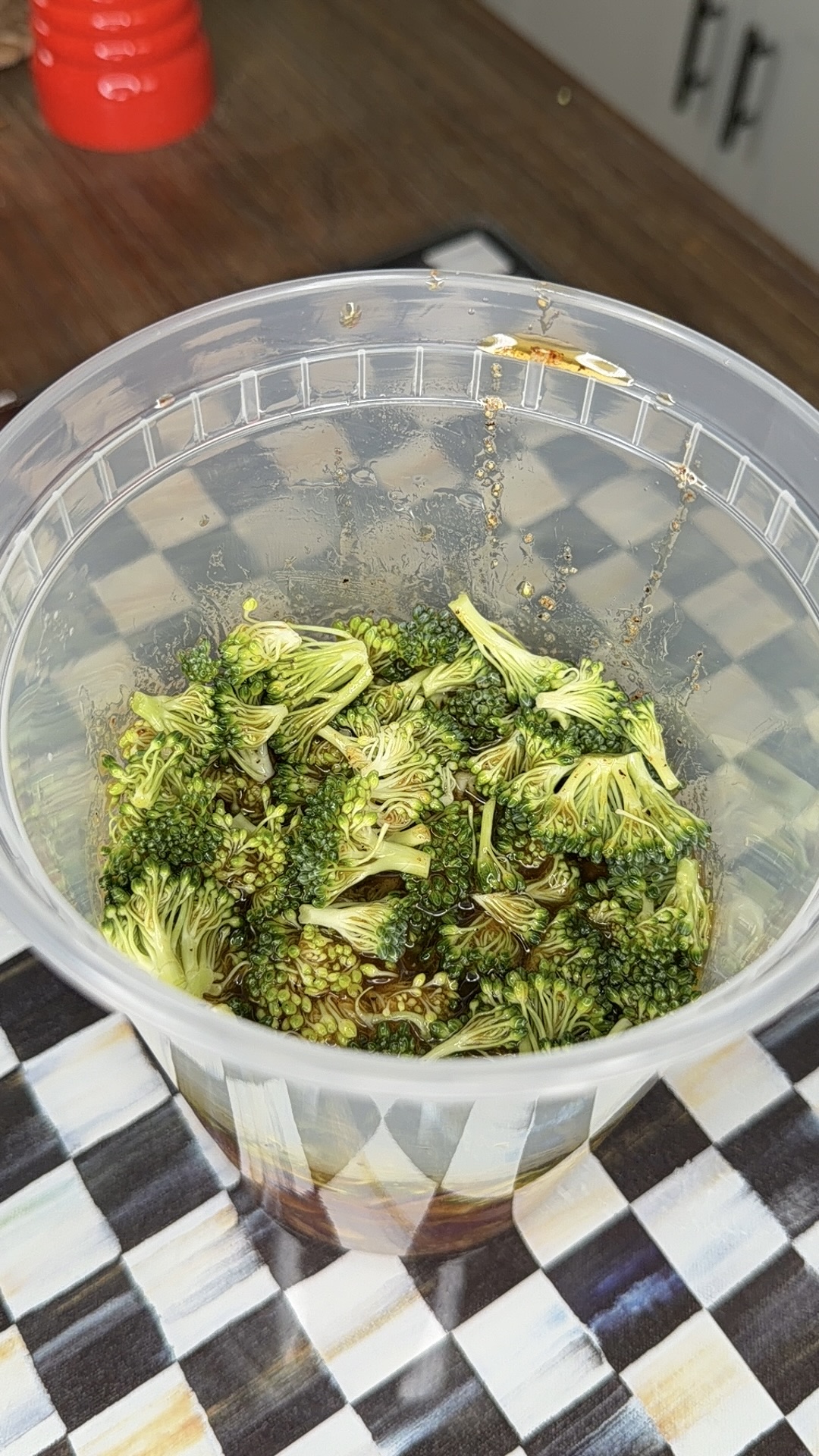 Chopped broccoli florets in a clear plastic cup, mixed with a dark soy sauce marinade, placed on a checkered black and white cloth on a wooden table.