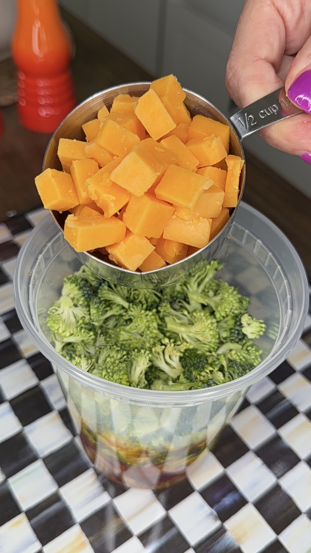 A hand holds a metal 1/2 cup measuring cup filled with diced cheddar cheese over a plastic container of chopped broccoli. The container sits on a checkered surface.