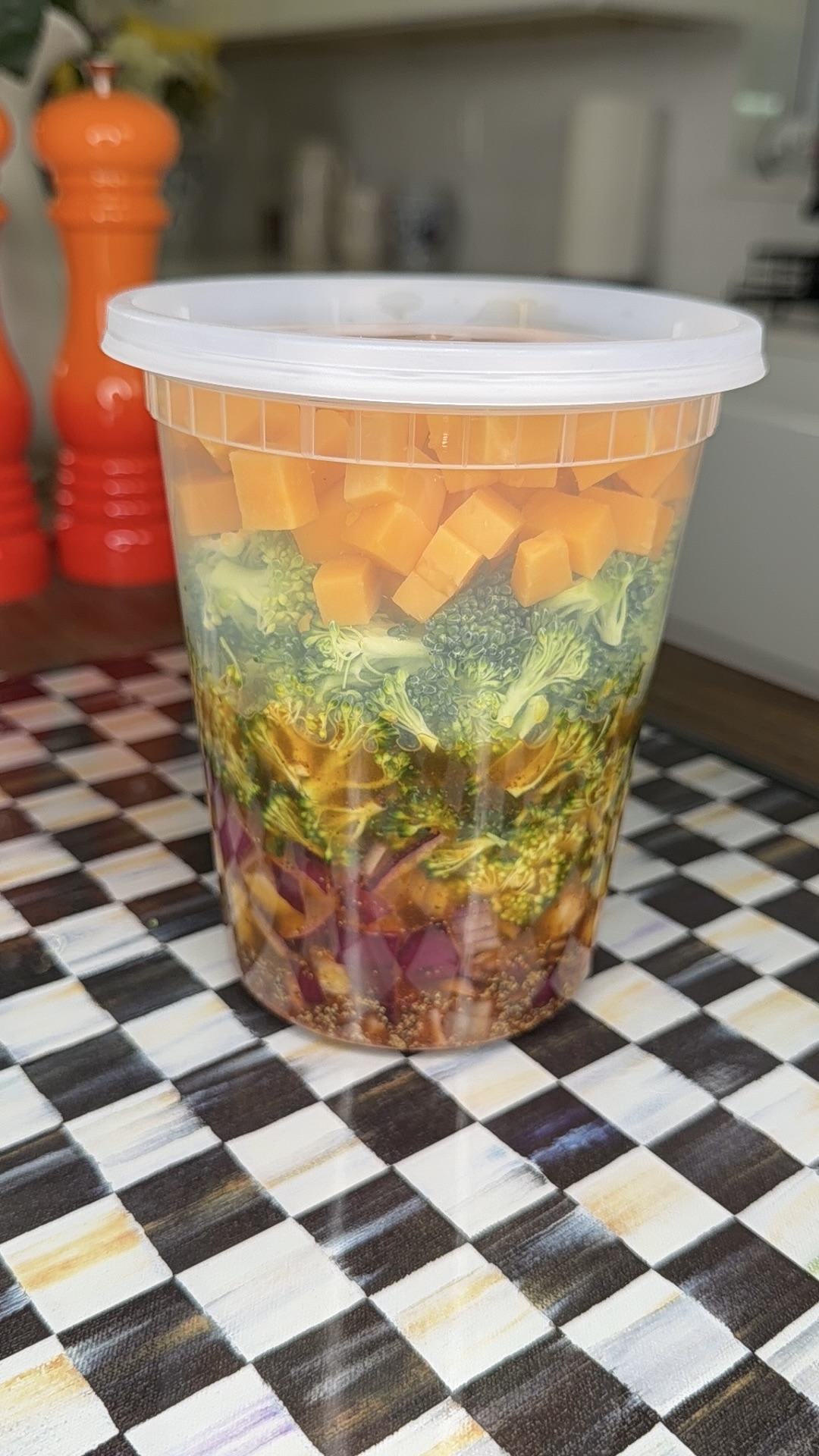 A clear plastic container filled with layered salad ingredients, including diced cheese, broccoli, red cabbage, and dressing at the bottom, sits on a checkered surface with orange condiment shakers in the background.