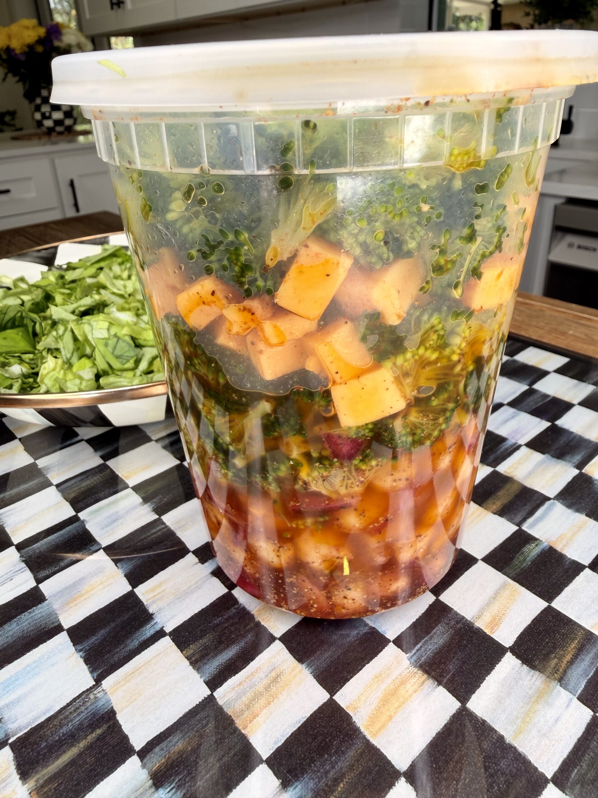 A large plastic container filled with a colorful vegetable and cheese salad in a spicy-looking dressing sits on a checkered tablecloth, with chopped greens on a plate nearby.