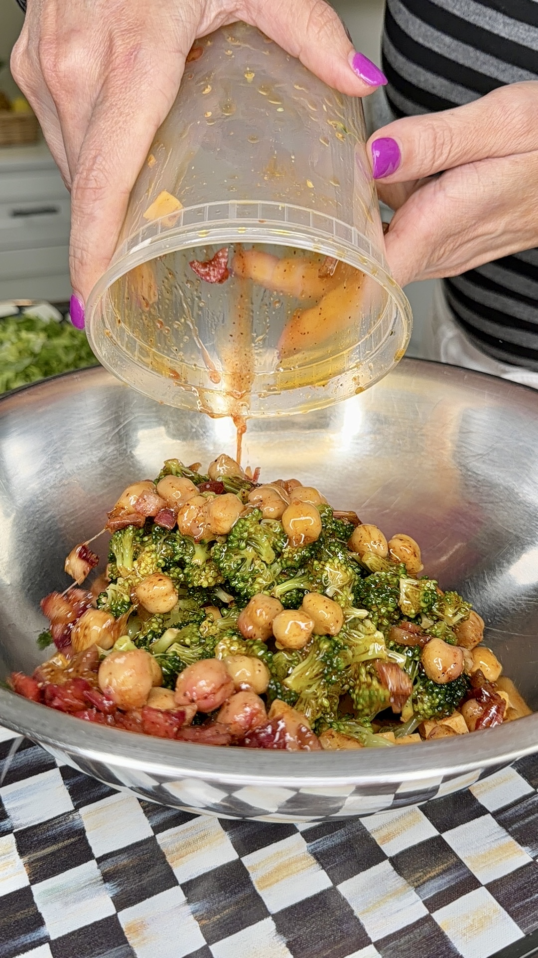 A person with pink nails pours dressing from a plastic container onto a salad with broccoli, chickpeas, and sun-dried tomatoes in a large metal bowl on a patterned tablecloth.