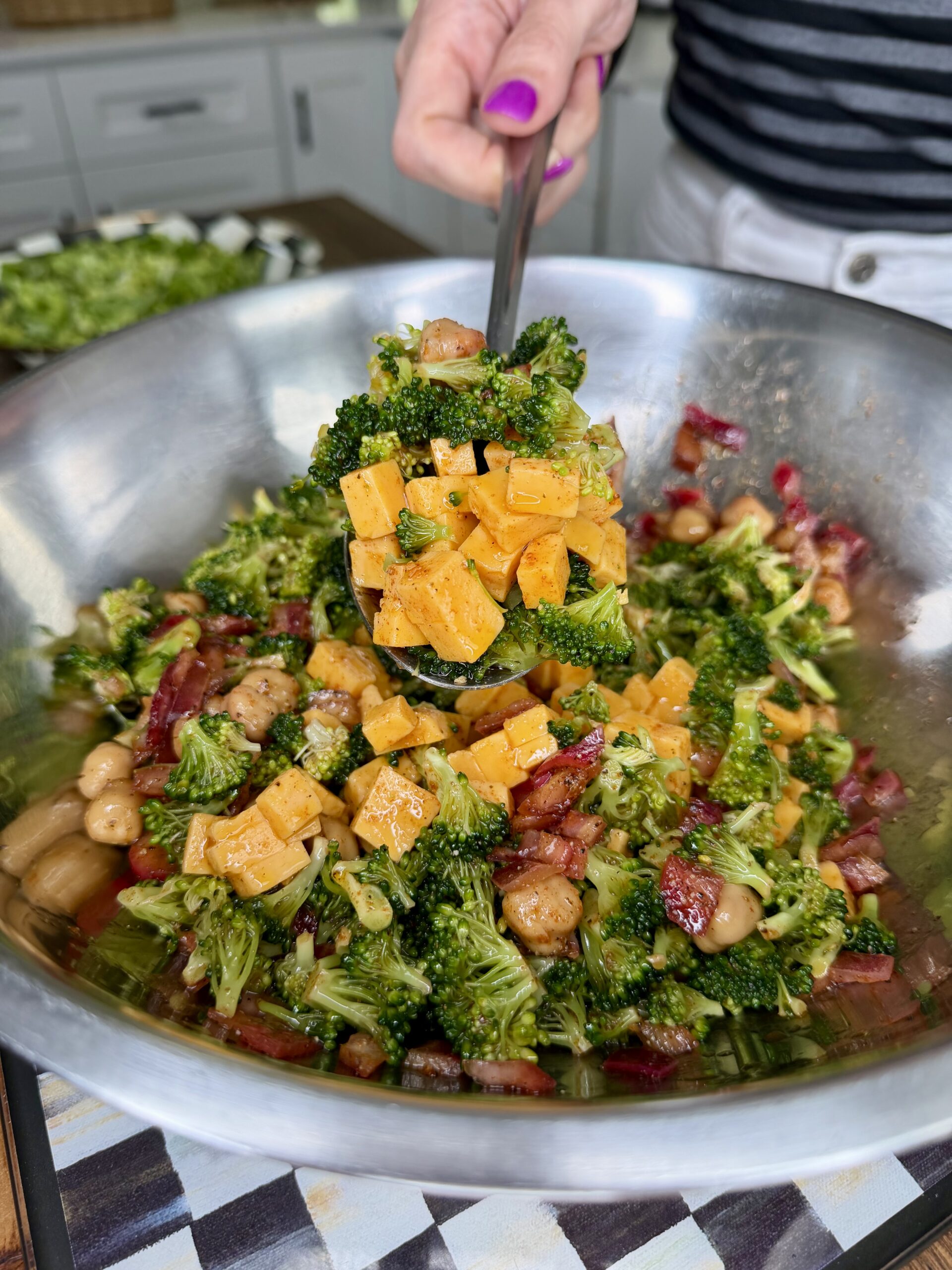 A close-up of a spoon lifting a serving of broccoli salad with cheddar cheese cubes, bacon, and beans from a large metal mixing bowl. A person with purple-painted nails holds the spoon.