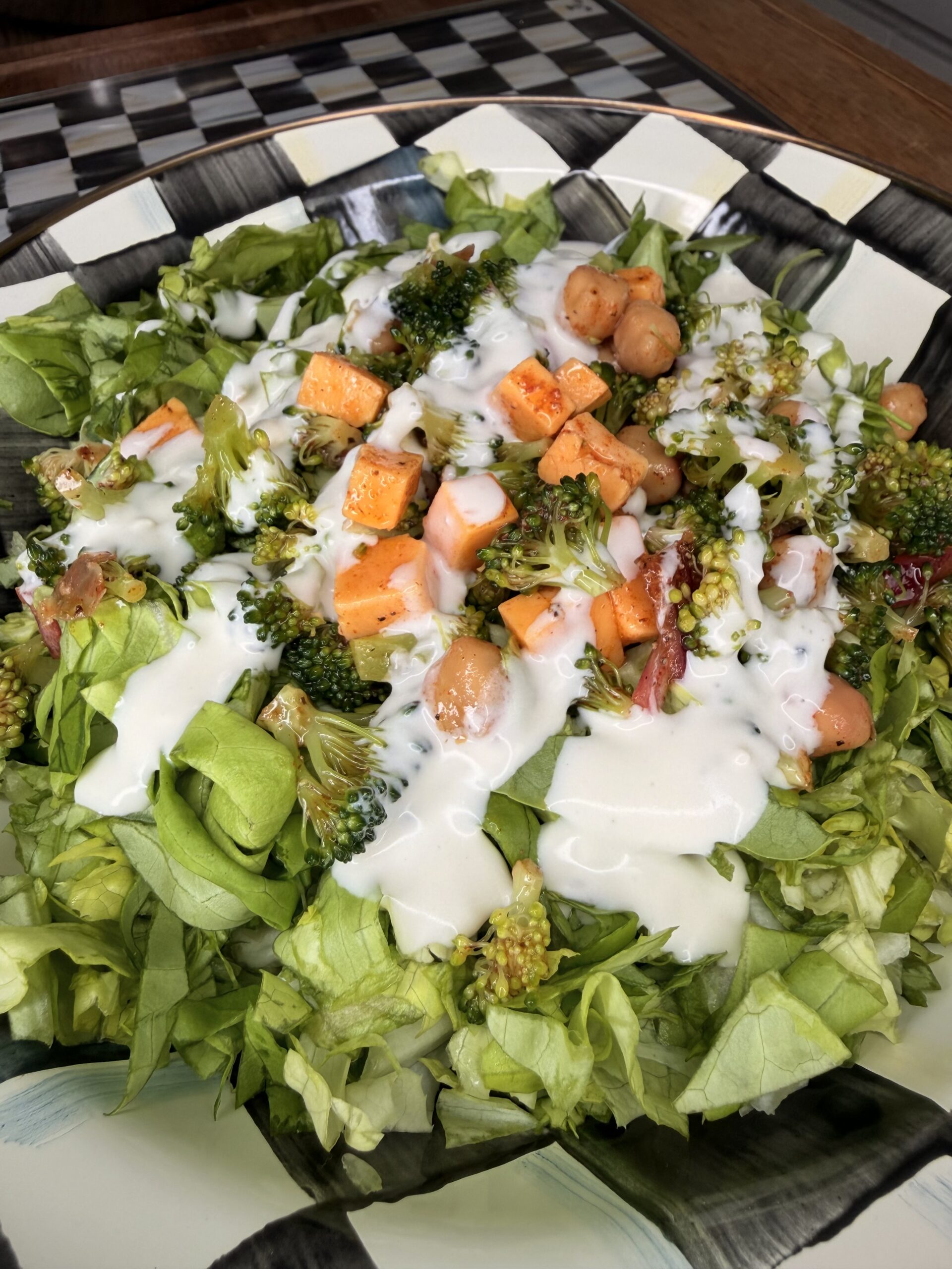 A bowl of fresh salad with chopped lettuce, broccoli, chickpeas, diced sweet potatoes, and creamy white dressing drizzled on top, served in a black and white patterned bowl.