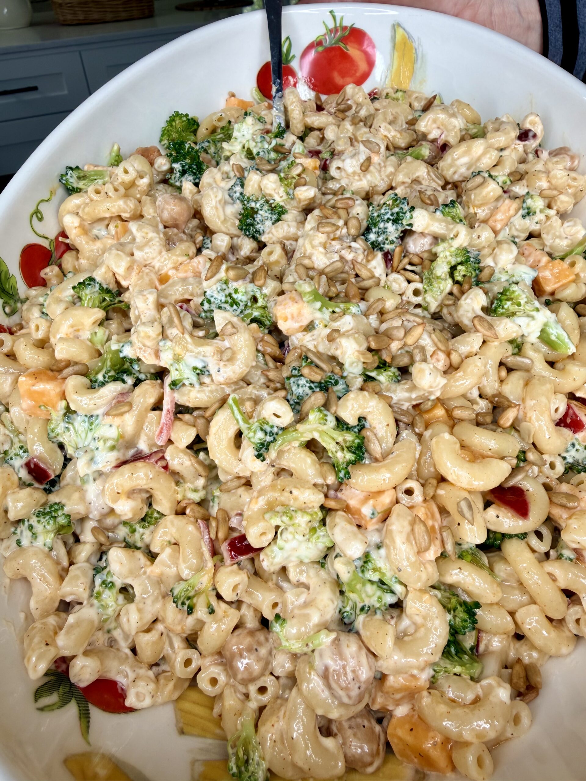 A close-up of a bowl filled with creamy pasta salad, containing elbow macaroni, broccoli, cherry tomatoes, sunflower seeds, and dressing, with a serving spoon partially visible.