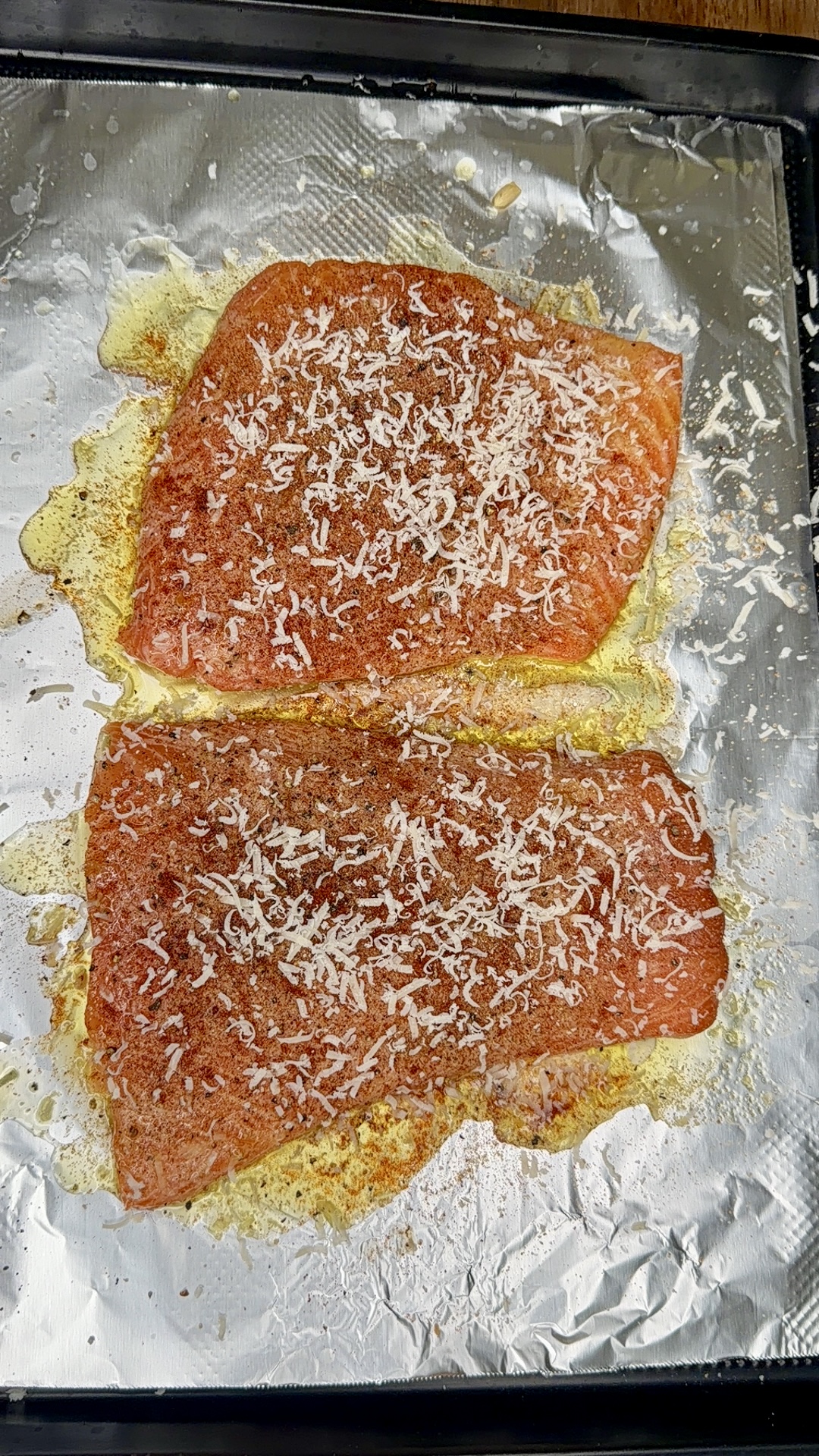 Two seasoned salmon fillets topped with grated cheese are baking on a foil-lined tray. The fish is golden and slightly crispy around the edges, with melted cheese sprinkled evenly on top.