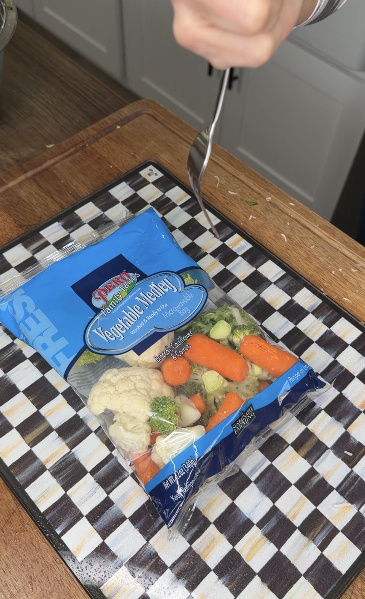 A hand holding a fork is about to open a blue bag of fresh vegetable medley containing carrots, cauliflower, and broccoli on a checkered cutting board in a kitchen.