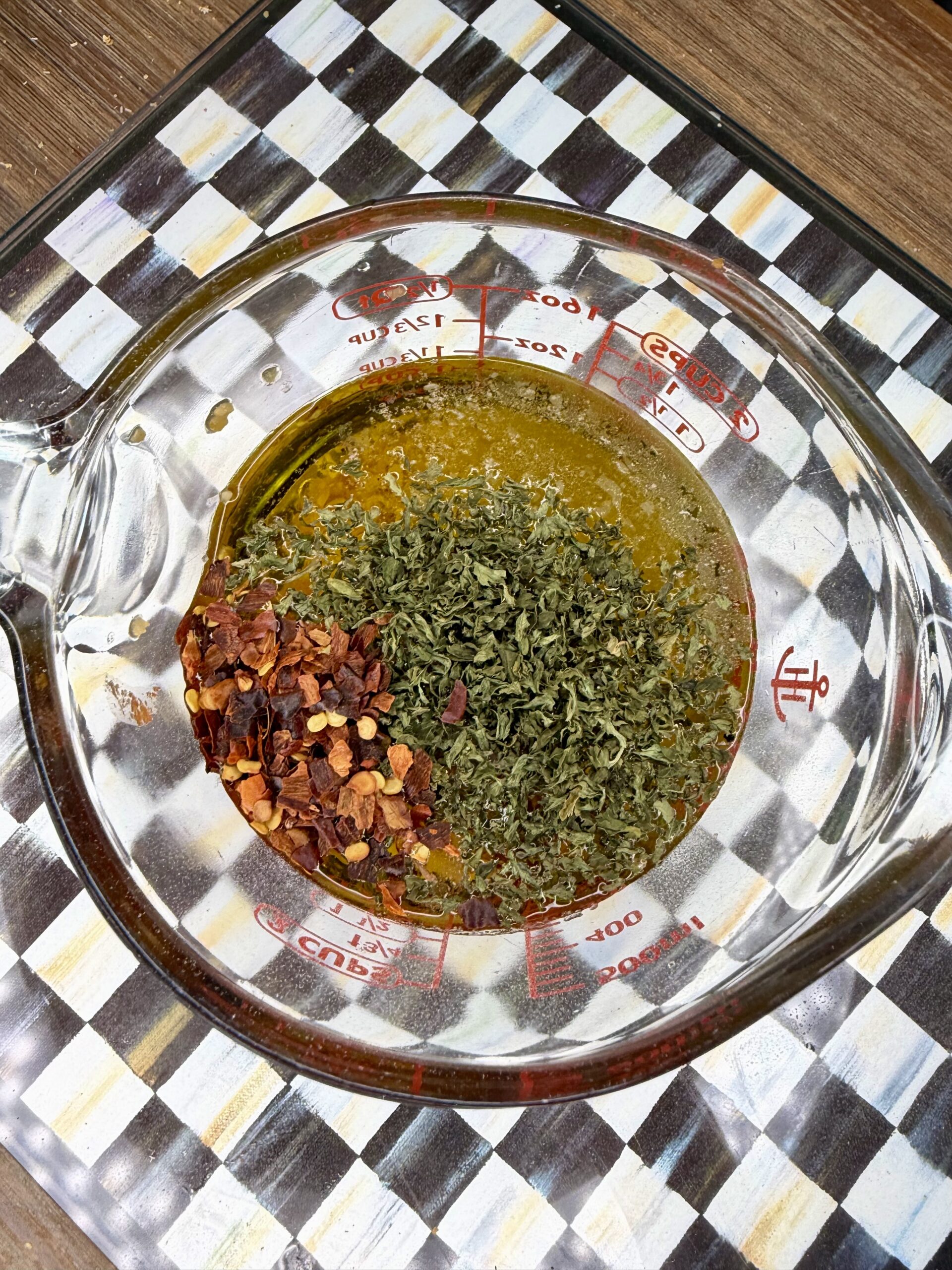 A glass measuring cup contains oil, dried parsley, and red pepper flakes, sitting on a checkered surface. The ingredients are unmixed and visible from above.