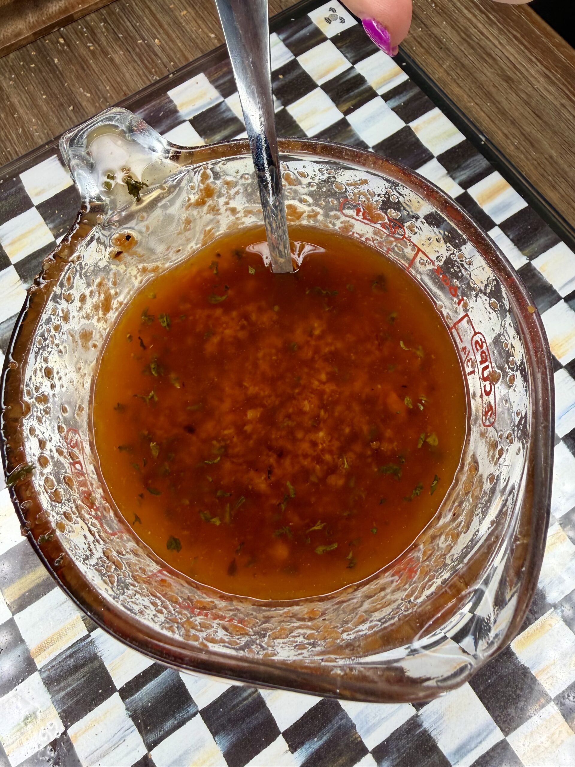A glass measuring cup filled with a reddish-brown liquid sauce mixed with herbs and minced garlic, with a metal spoon inside. The cup sits on a patterned surface, with a hand holding the spoon.