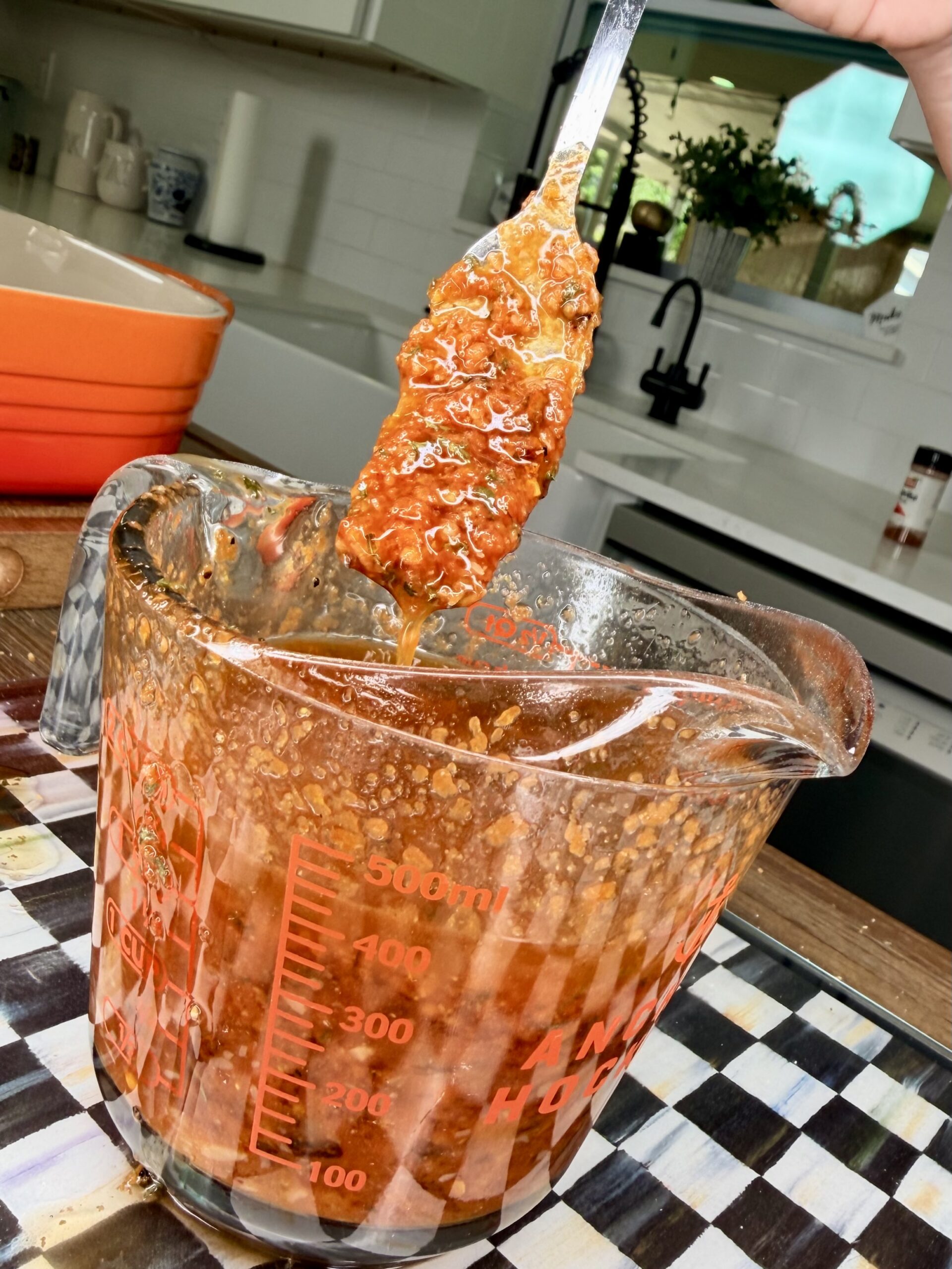 A hand holds a spoon lifting a piece of food coated in a thick, reddish-brown sauce from a glass measuring cup, with a kitchen setting visible in the background.
