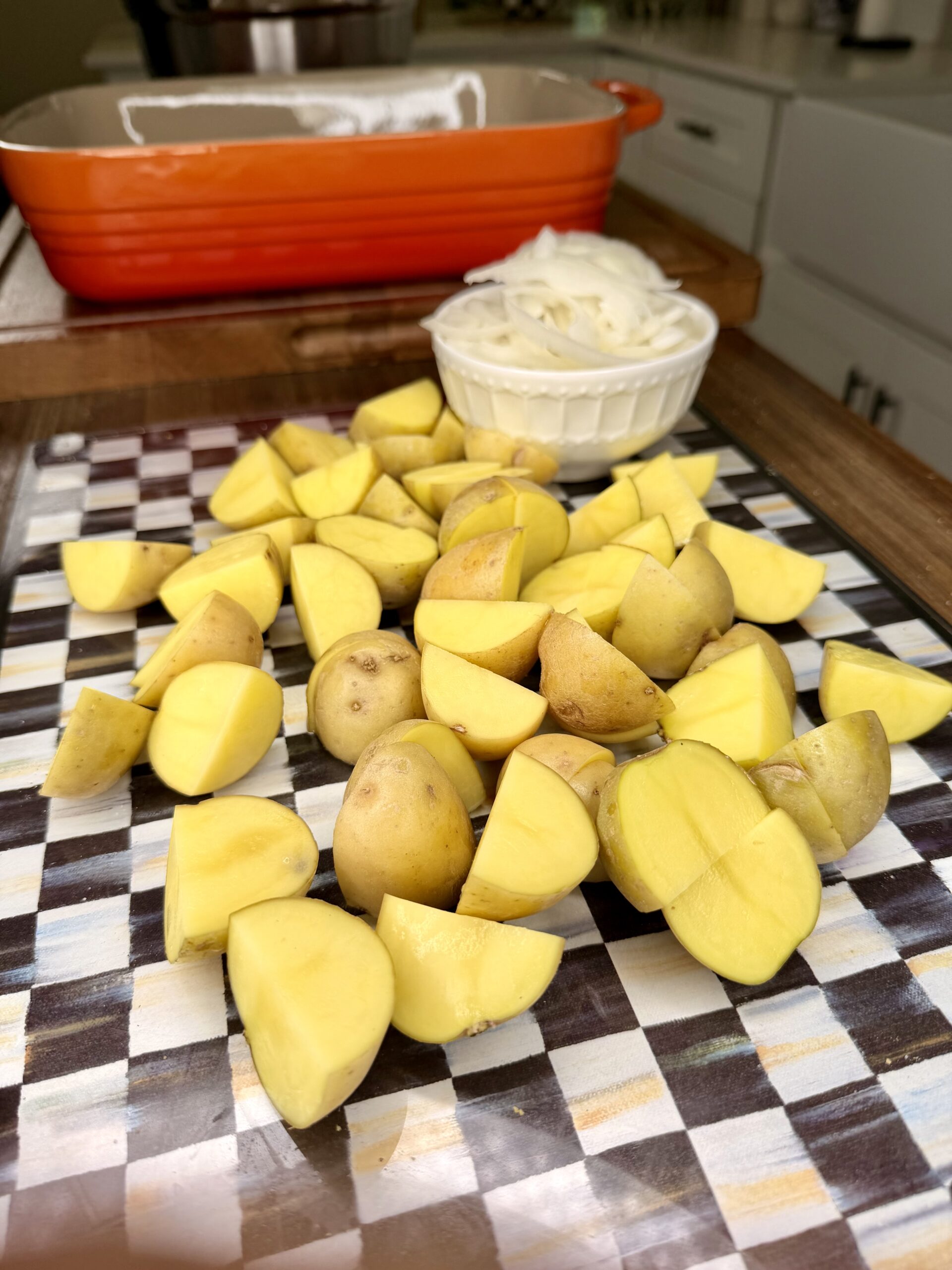 Chopped yellow potatoes are spread on a checkered cutting board. Behind them is a small white bowl filled with sliced onions and an orange baking dish on a kitchen countertop.
