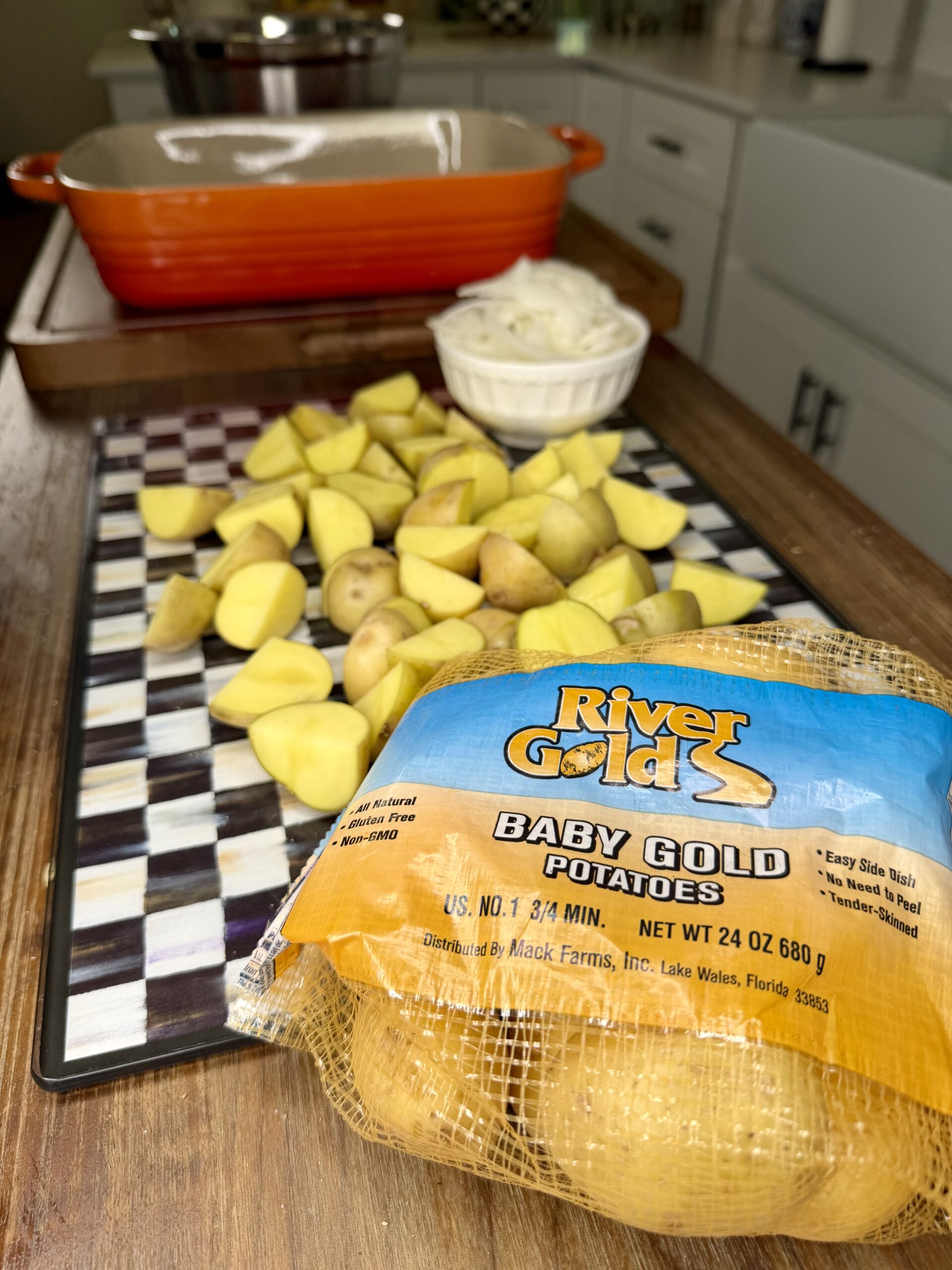 A bag of River Gold baby gold potatoes sits on a counter next to chopped potatoes on a checkered cutting board, with sliced onions in a small dish and an orange baking dish in the background.