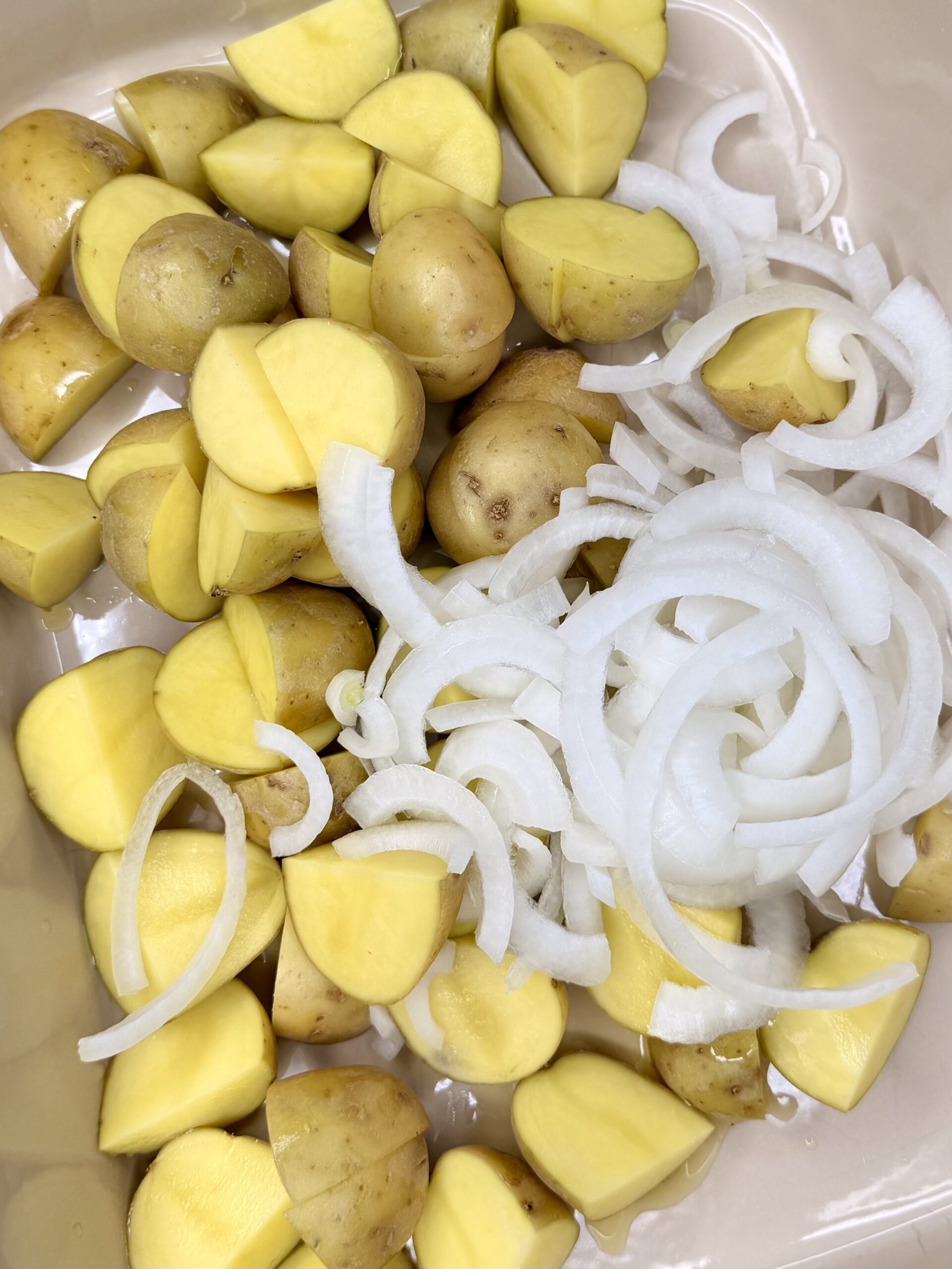 Quartered yellow potatoes and sliced white onions arranged together in a clear container, ready for cooking or roasting.