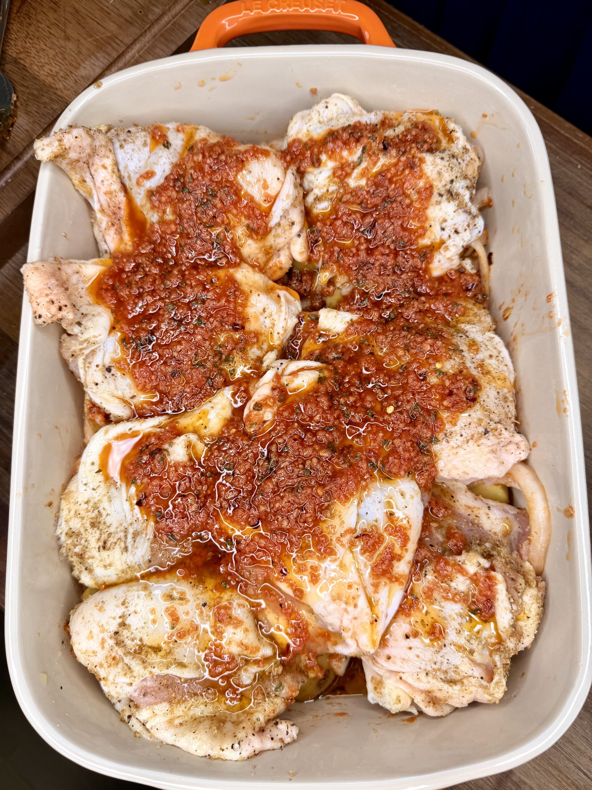 Raw chicken pieces in a baking dish are topped with a chunky, red marinade or sauce, likely seasoned with spices and herbs, ready to be cooked. The dish has an orange handle visible at the top.