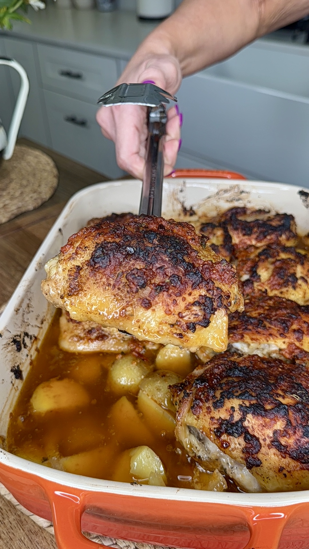 A hand uses tongs to lift a piece of crispy, browned chicken from a baking dish filled with roasted chicken thighs, potatoes, and flavorful juices. The dish sits on a wooden countertop in a kitchen.