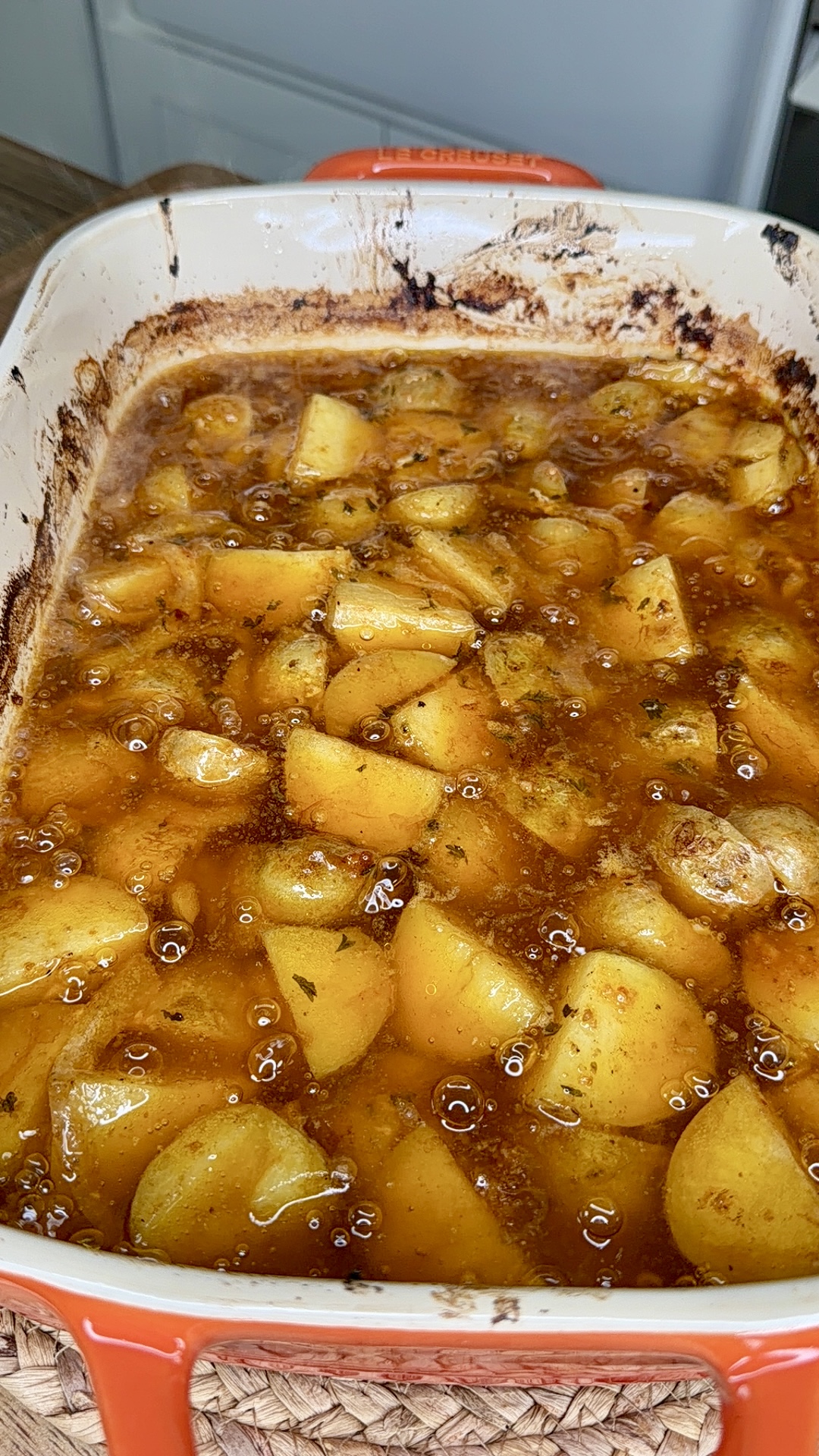 A close-up of a baking dish filled with golden, roasted potatoes coated in a glistening brown sauce and herbs, with some caramelization on the edges of the dish.