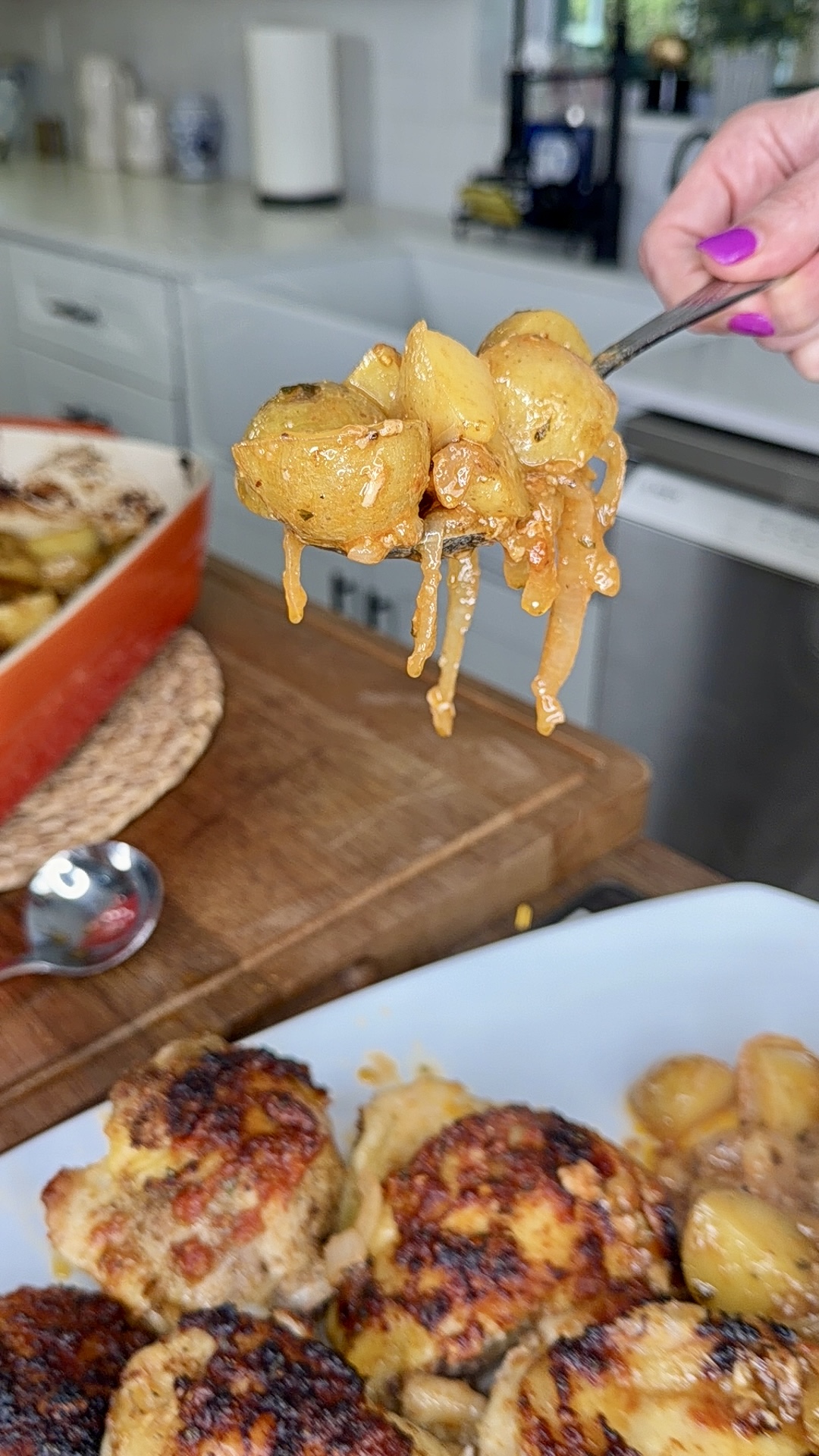 A close-up of a hand holding a fork with caramelized onions and roasted potatoes above a plate of browned meatballs and more potatoes in a kitchen setting.