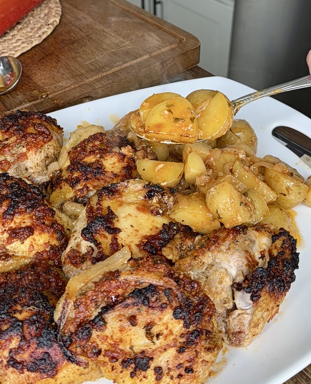 A plate of golden-brown roasted chicken pieces with a spoon serving cooked potatoes and onions on top. The food is arranged on a white platter on a kitchen counter.