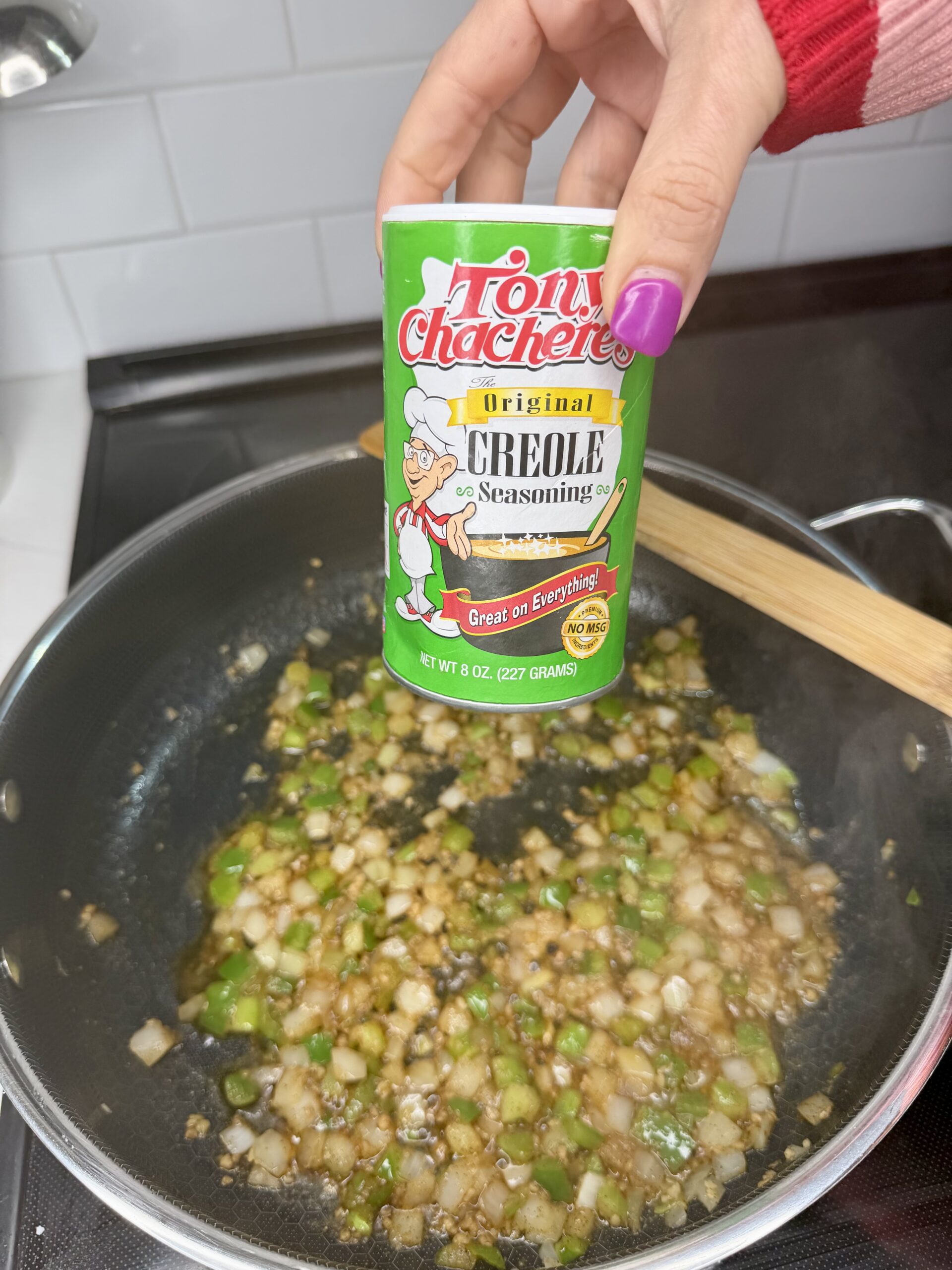 A hand holds a container of Tony Chacheres Creole Seasoning over a pan of chopped onions and green peppers cooking on a stovetop. A wooden spoon rests on the pan’s edge.