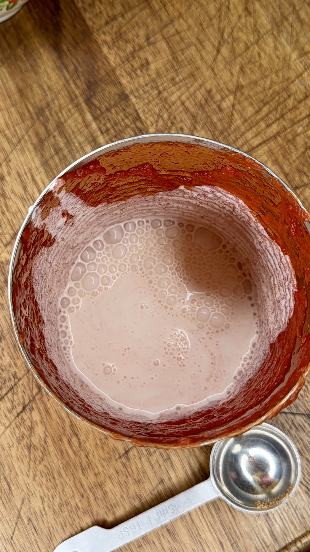 A metal mixing bowl with a pink, frothy liquid inside, and red residue on the sides, sits on a wooden surface next to a white measuring spoon and a metal measuring scoop.