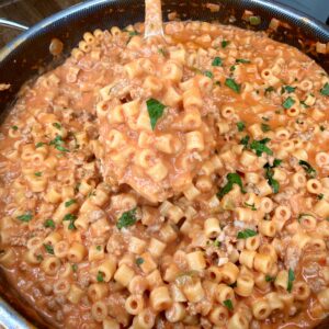 A skillet filled with creamy tomato pasta featuring small tube-shaped noodles, ground meat, and garnished with fresh chopped herbs. A ladle is lifting a portion from the pan.