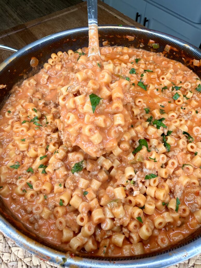 A skillet filled with creamy tomato pasta featuring small tube-shaped noodles, ground meat, and garnished with fresh chopped herbs. A ladle is lifting a portion from the pan.