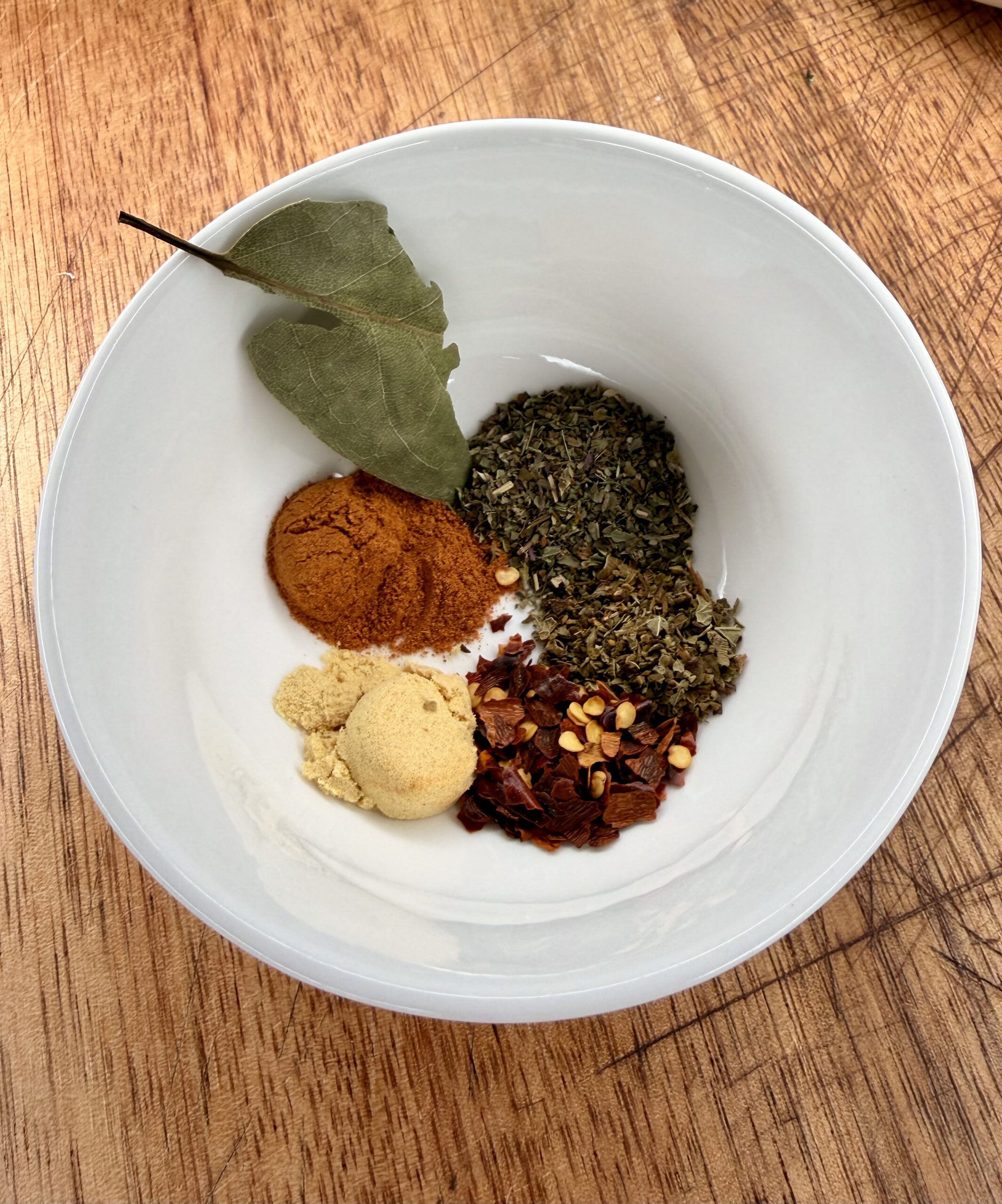 A white bowl on a wooden surface containing a bay leaf, ground turmeric, ground ginger, dried basil, crushed red pepper flakes, and dried chili flakes arranged in small piles.