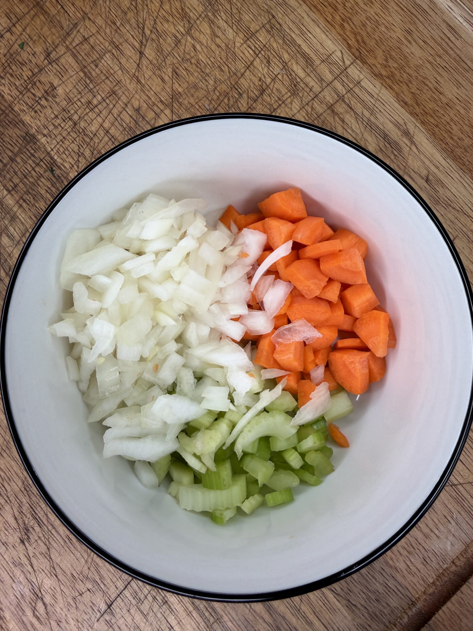 A white bowl on a wooden surface contains chopped onions, carrots, and celery, arranged in separate sections.