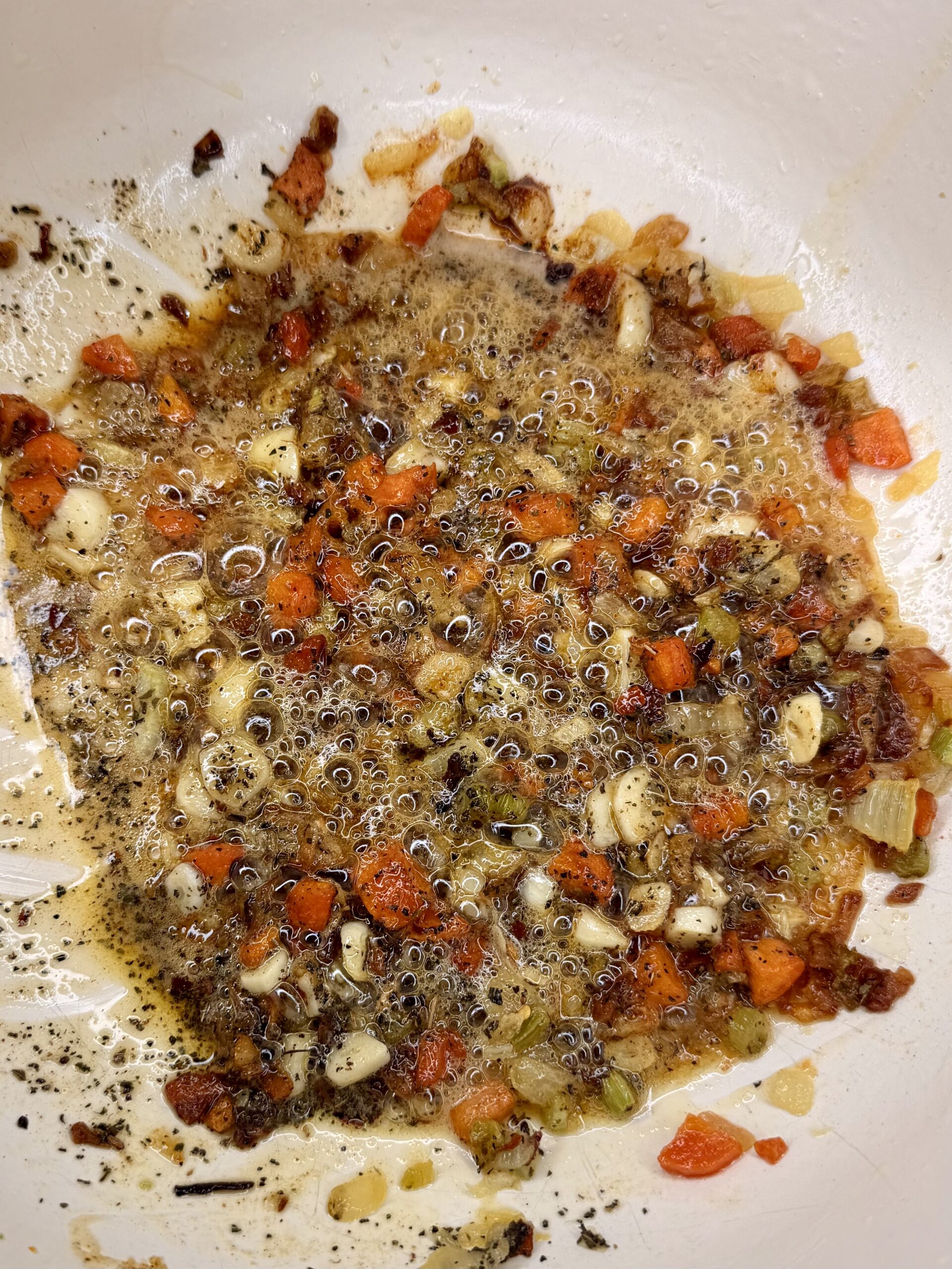 Chopped onions, garlic, and red bell peppers sautéing in bubbling oil and seasonings in a white pan, with some browned and caramelized bits throughout.