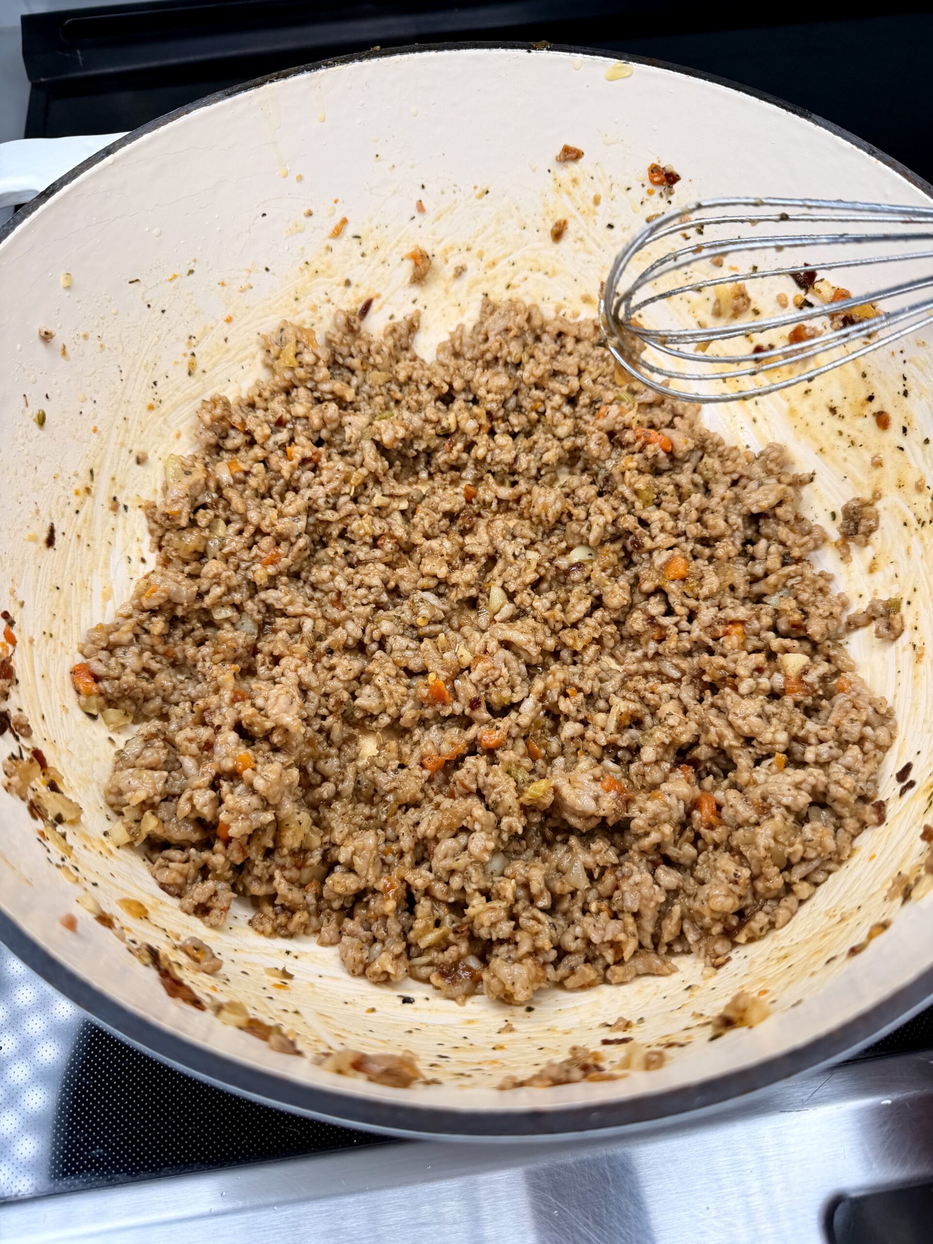 Cooked ground meat mixed with finely chopped carrots, onions, and seasonings in a large white pot. A metal whisk rests on the side of the pot on a stovetop.