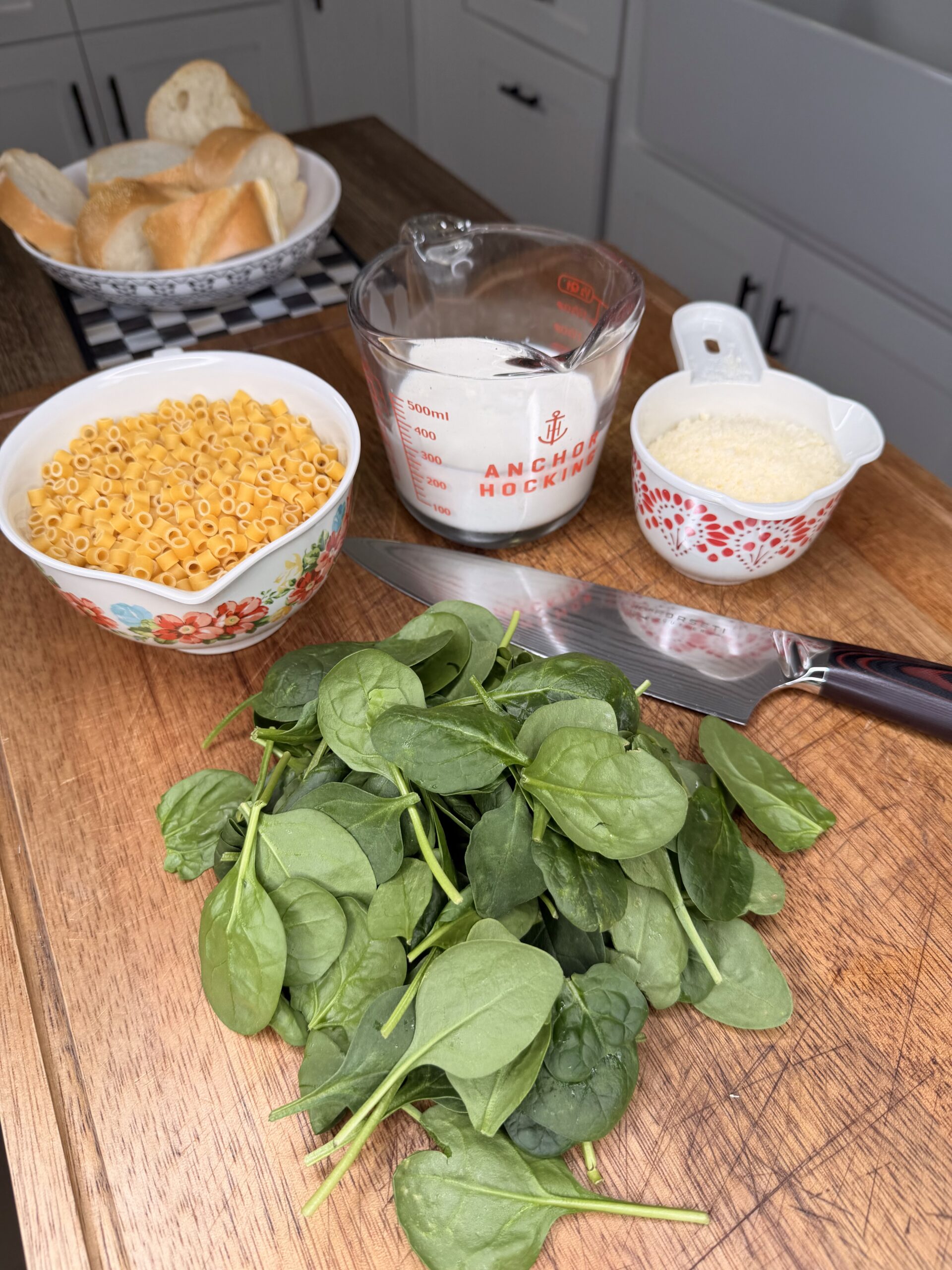 A wooden countertop displays fresh spinach, a bowl of dry pasta, a glass measuring cup with milk, a small bowl of grated parmesan, a knife, and a plate of sliced bread in the background.