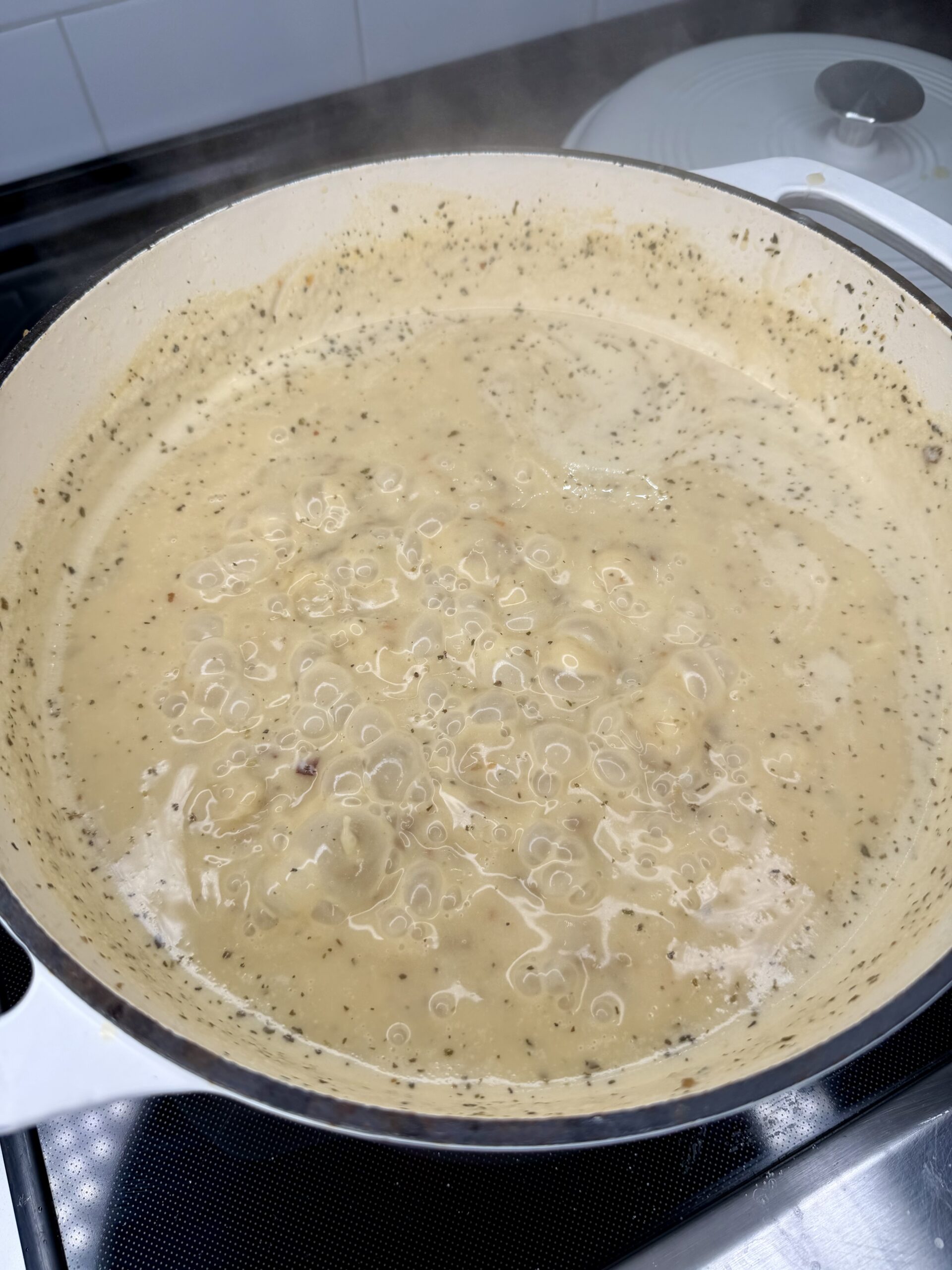 A creamy, beige sauce with herbs and visible bubbles simmers in a white pot on a stovetop. A white lid rests nearby, and the sauce appears thick and well-mixed.