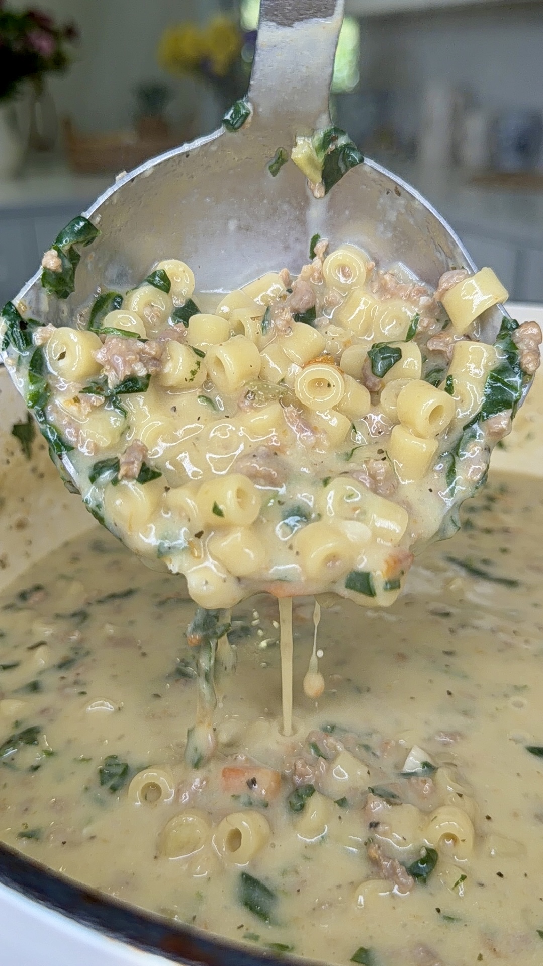 A close-up of a ladle lifting creamy macaroni soup with ground meat and spinach, with stretchy melted cheese visible and the pot seen below.