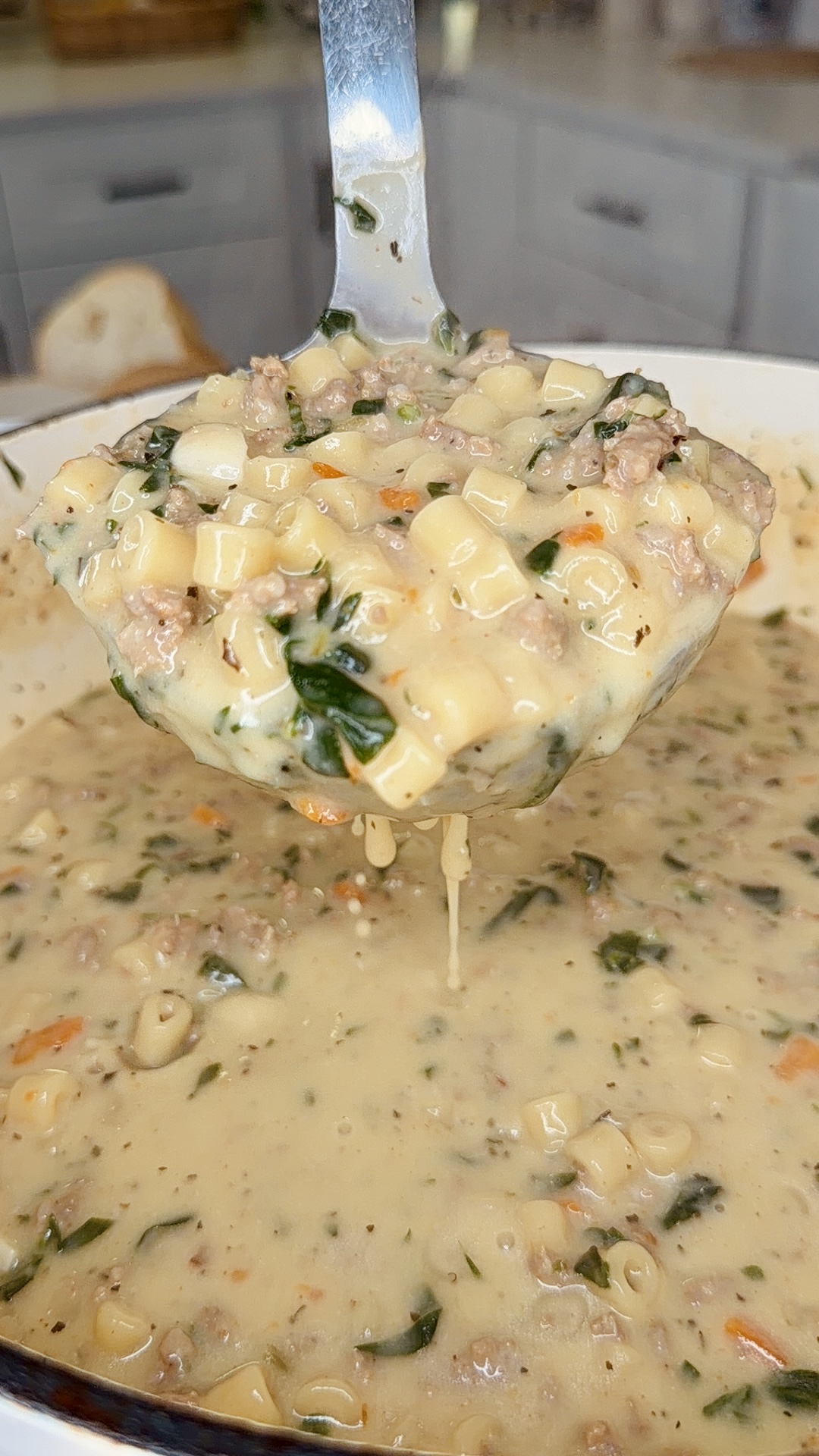 A close-up of a ladle lifting creamy soup filled with diced pasta, ground meat, spinach, and herbs from a pot in a kitchen.